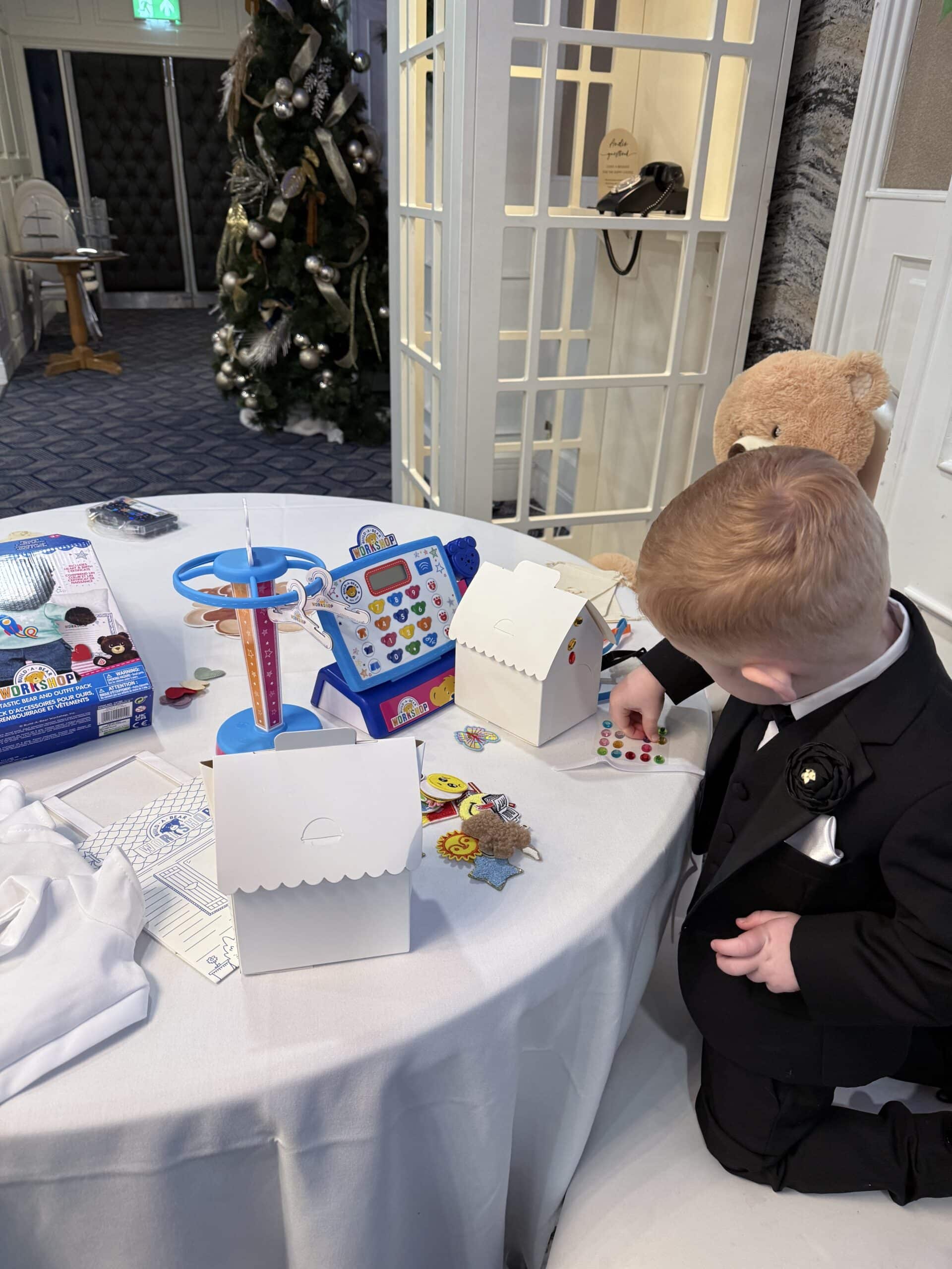 A child in a suit decorates a box with stickers at a table, with toys and a Christmas tree in the background.