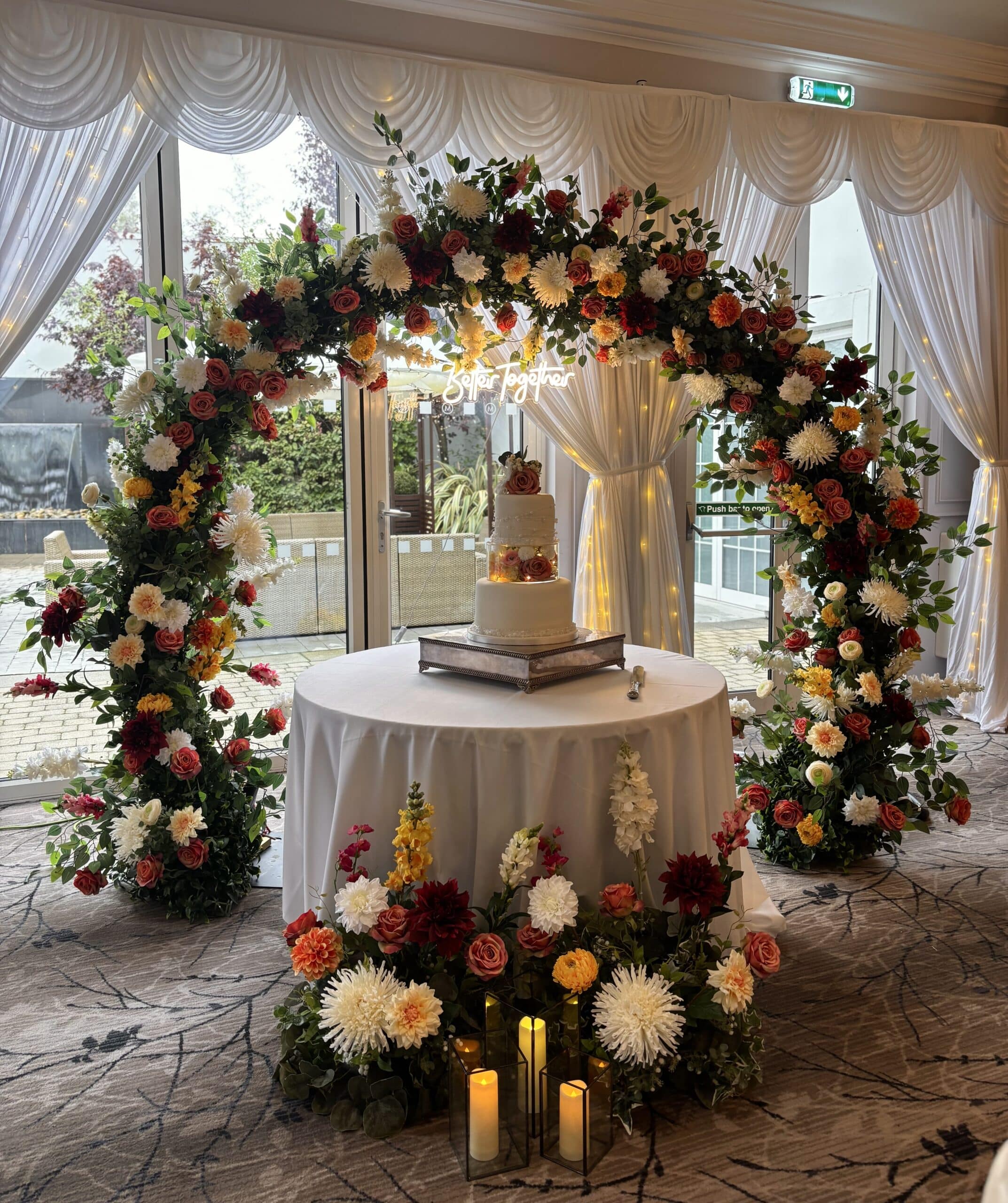 Wedding cake table with floral arch and candles in elegantly decorated room.