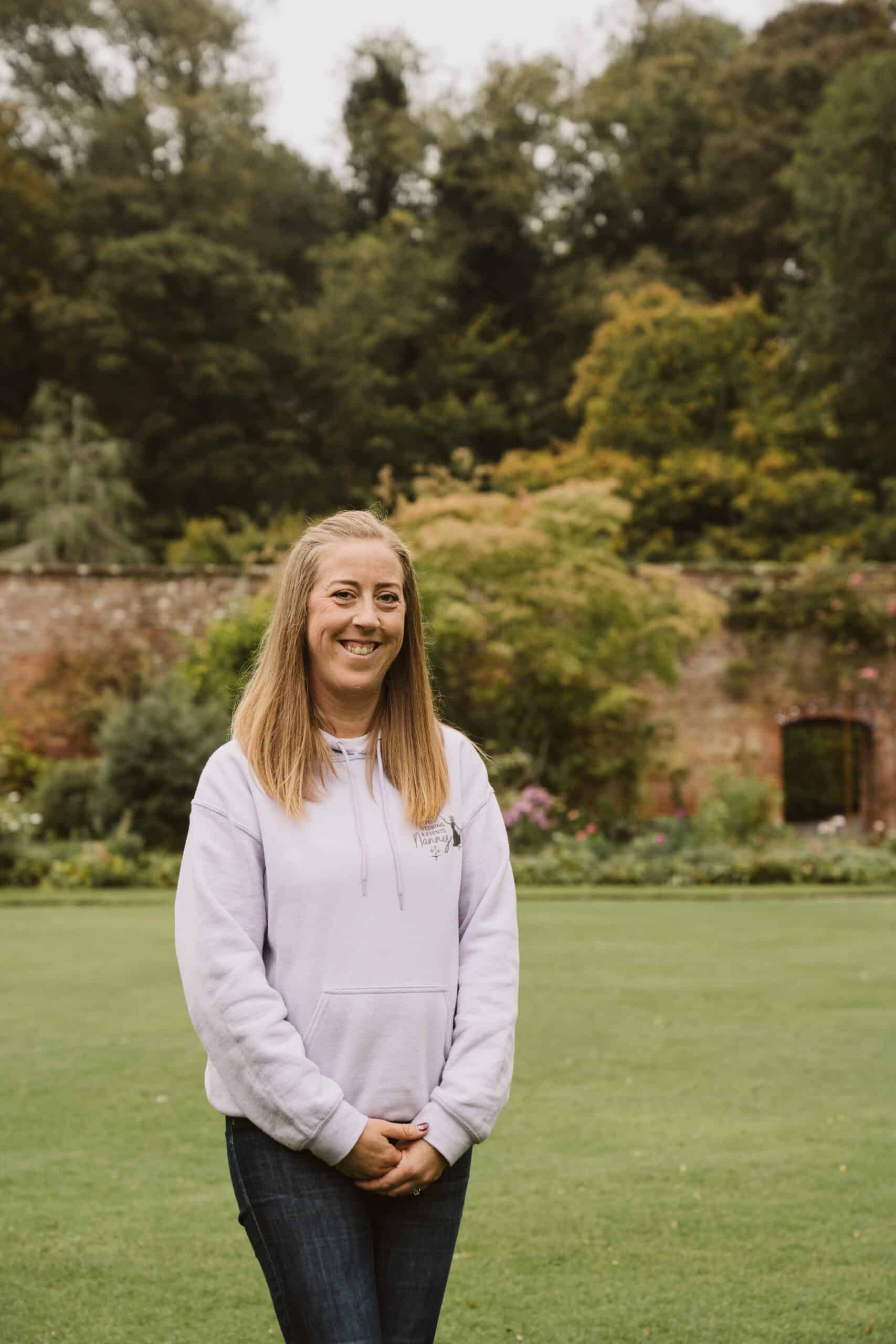 Woman in a light hoodie smiling in a garden with a stone wall and trees in the background.