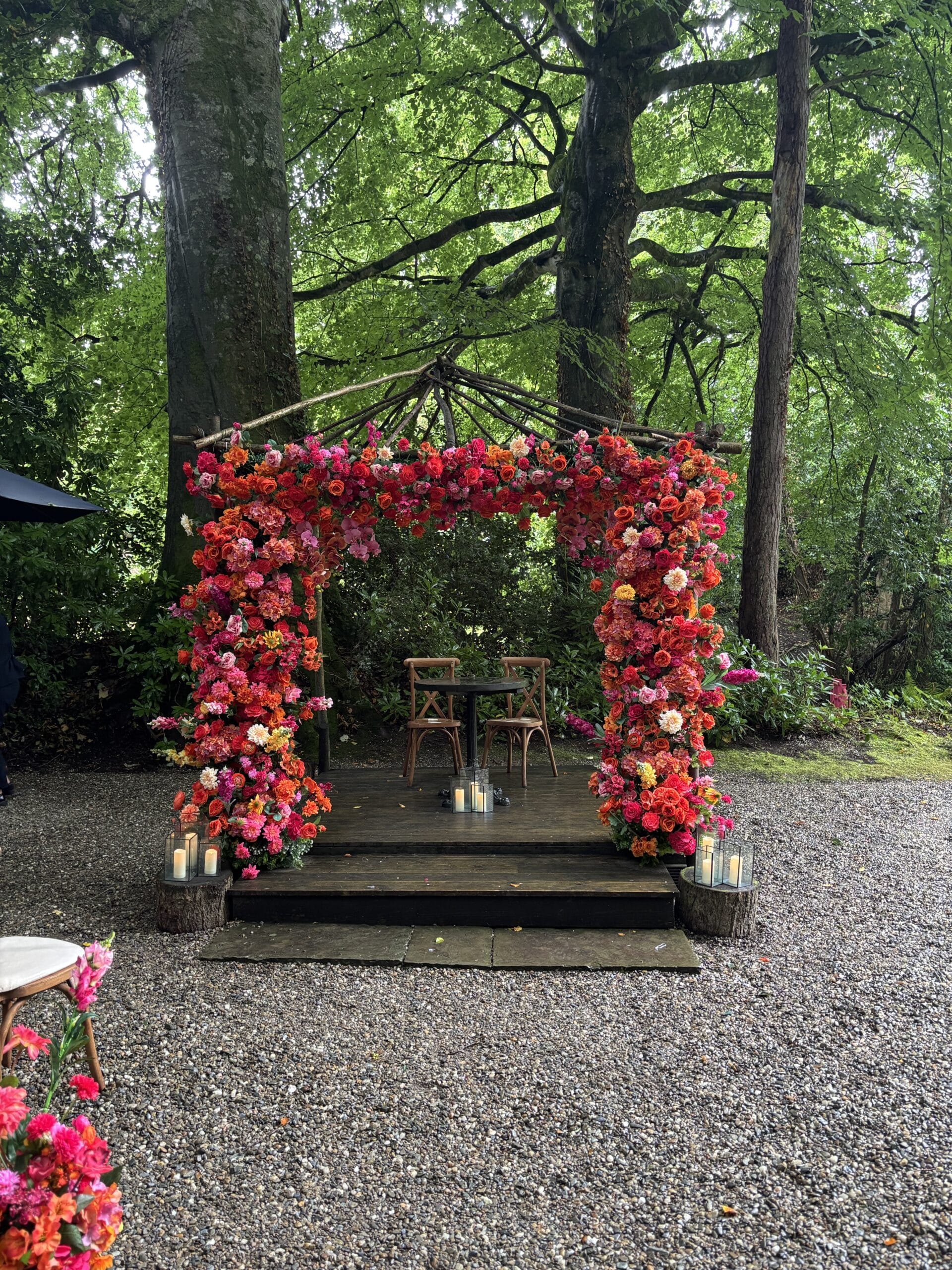 Flower-adorned outdoor wedding arch under trees with candles and chairs.