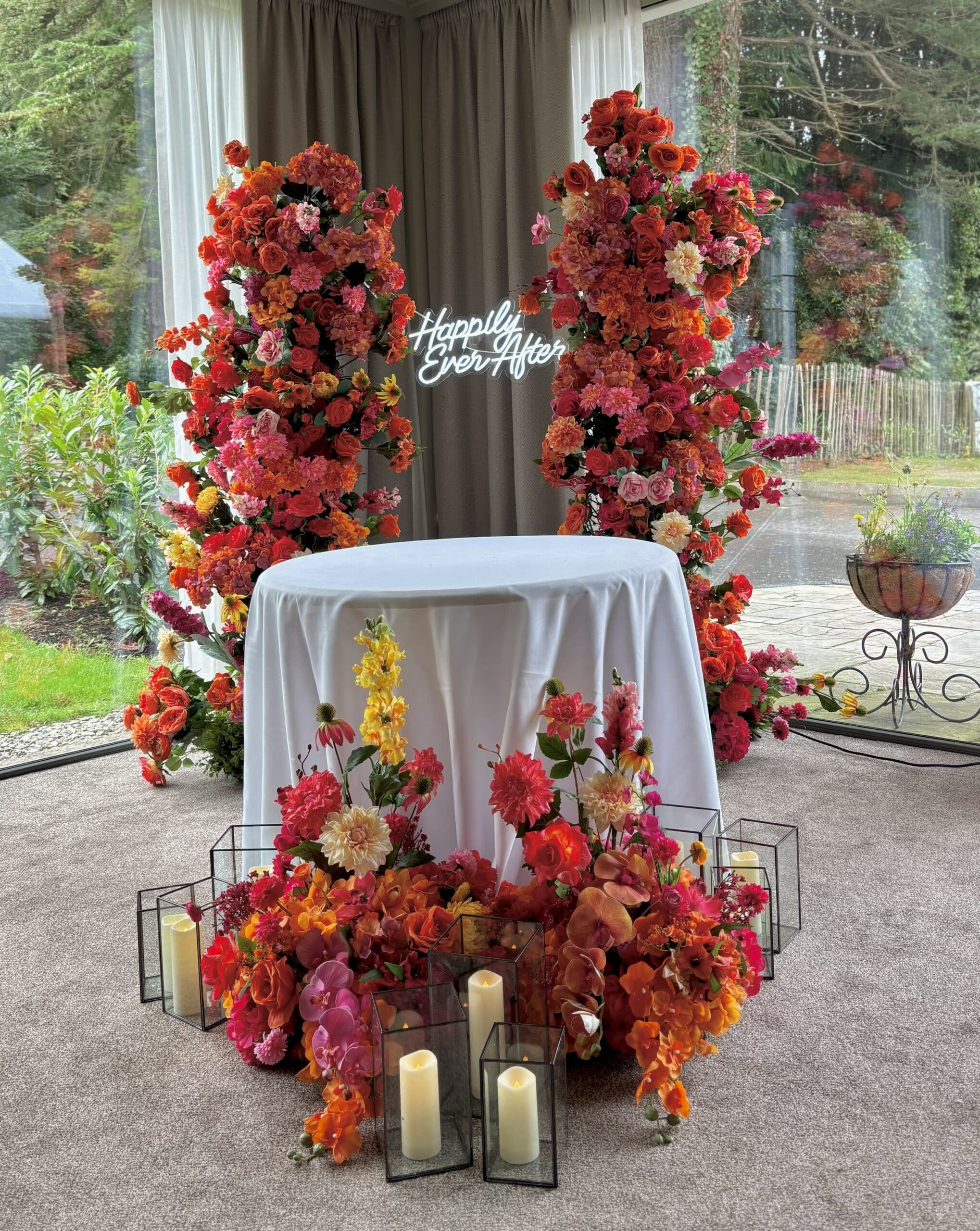 Wedding decor with vibrant floral arrangements, lit candles, and "Happily Ever After" neon sign behind a draped table.