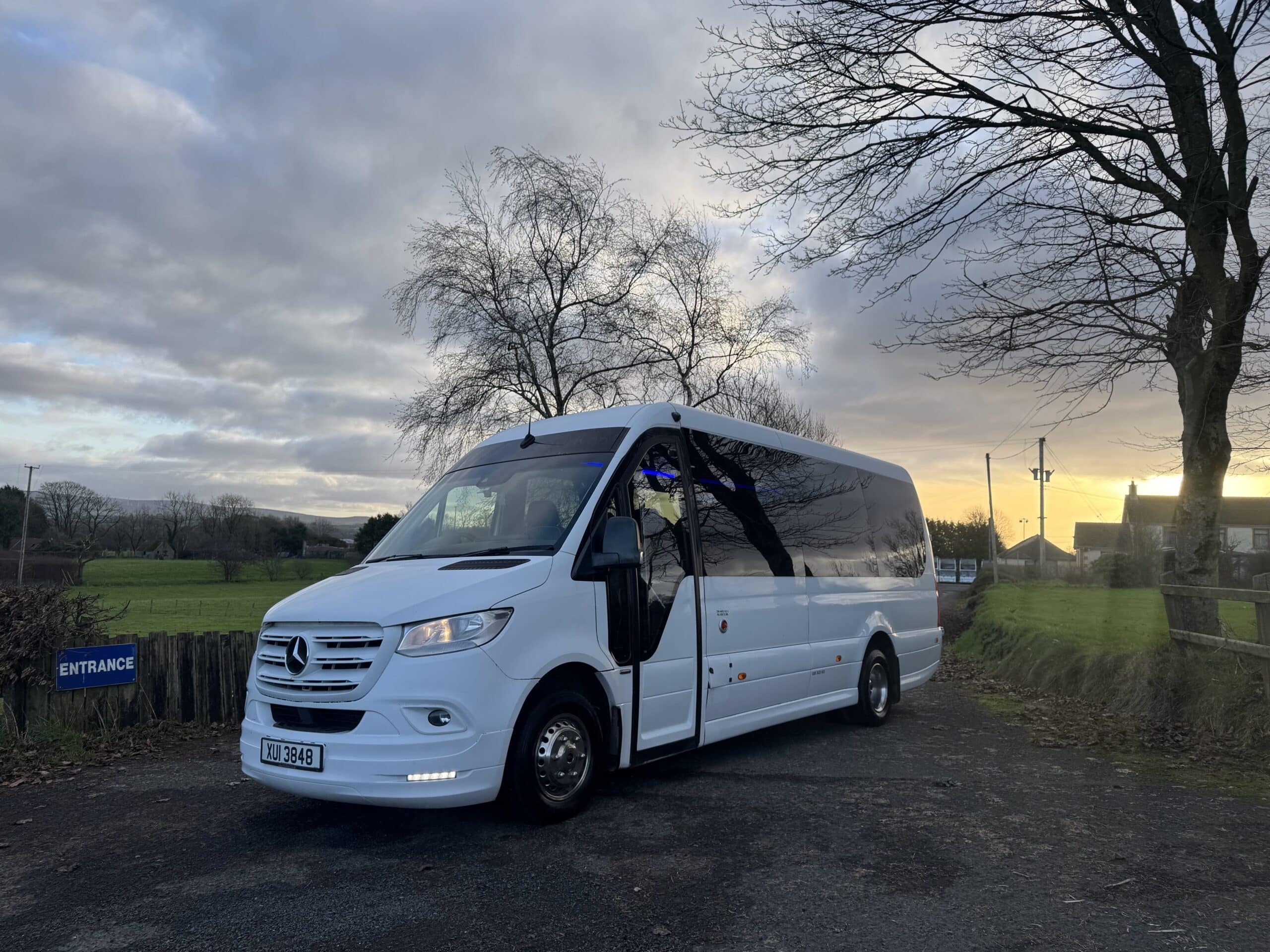 White mini-bus parked by rural entrance at sunset with trees and cloudy sky in the background.