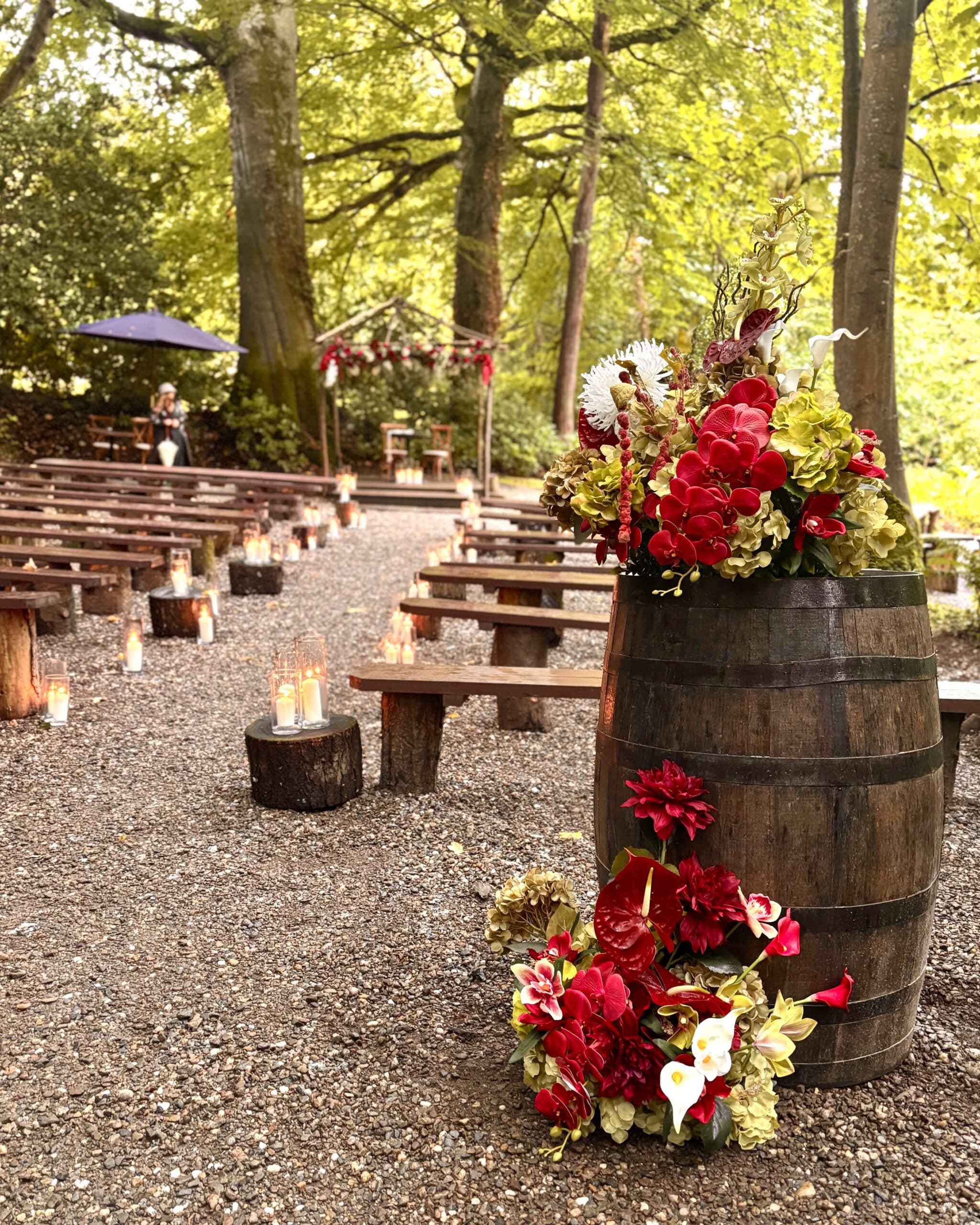 Outdoor wedding setup with floral arrangements, wooden benches, candles, and a rustic barrel in a forest setting.