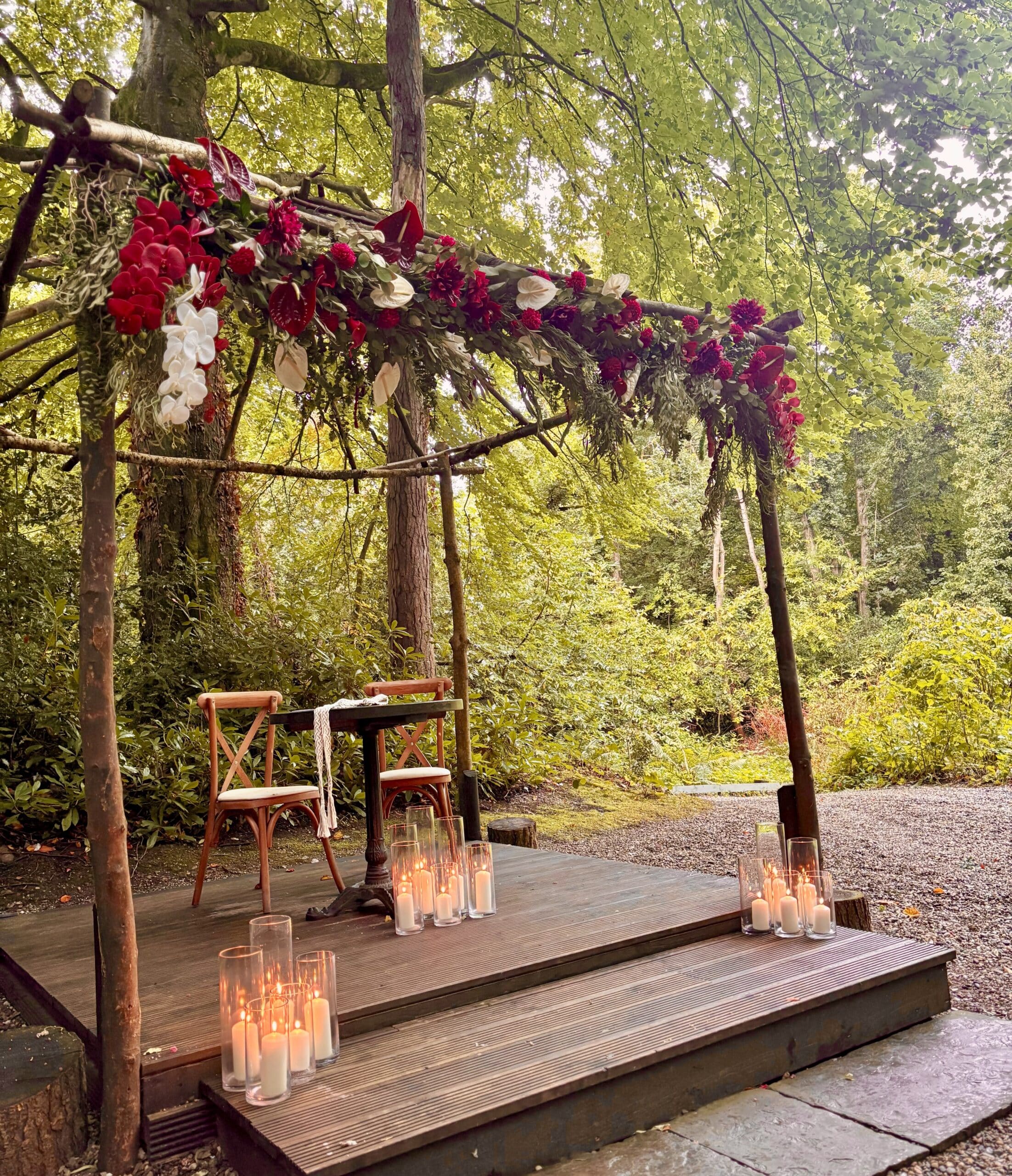 Outdoor wedding setup with a floral arch, wooden chairs, a table, and candles on a wooden platform in a forest setting.
