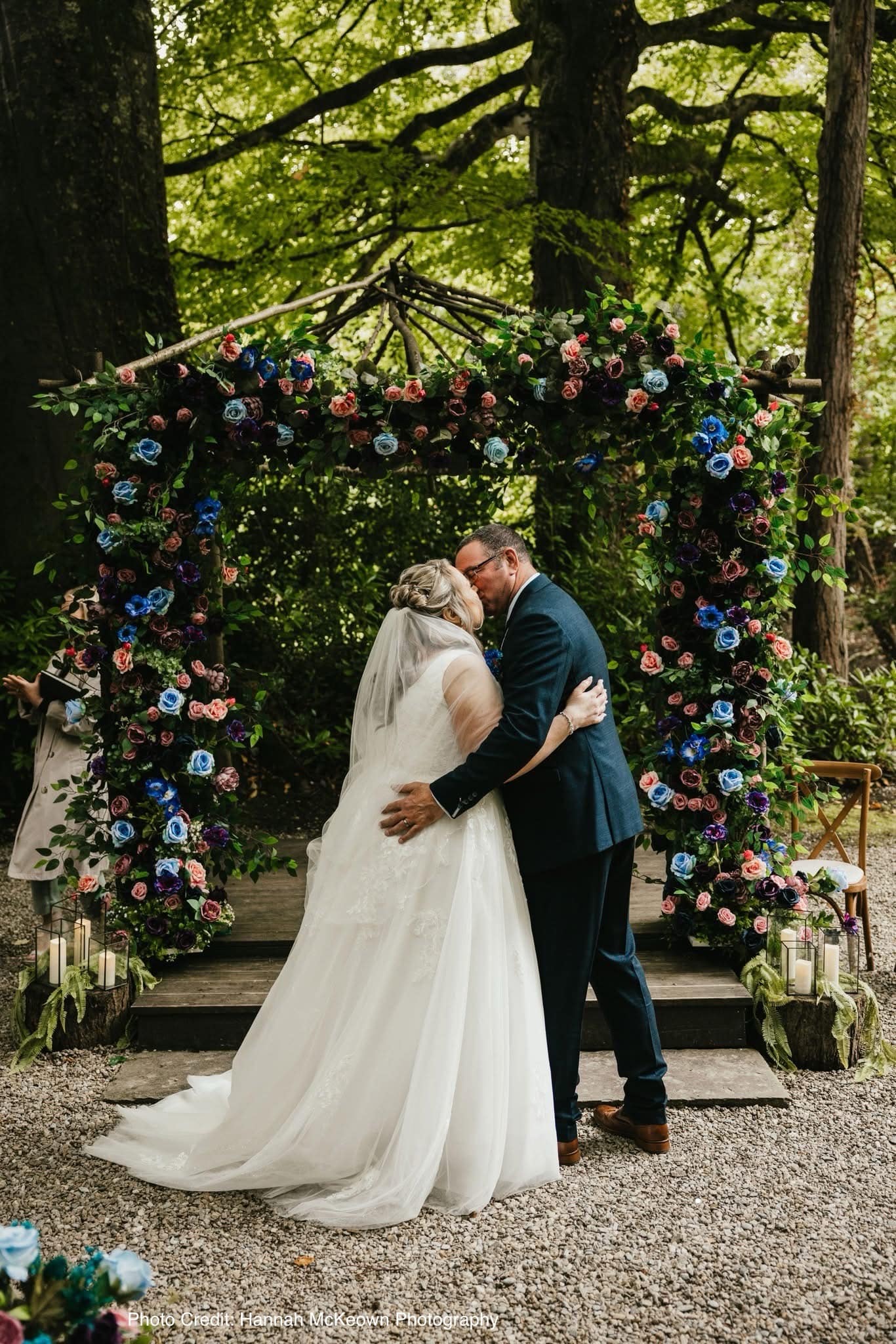 Bride and groom kissing under a colorful floral arch in an outdoor wedding ceremony.