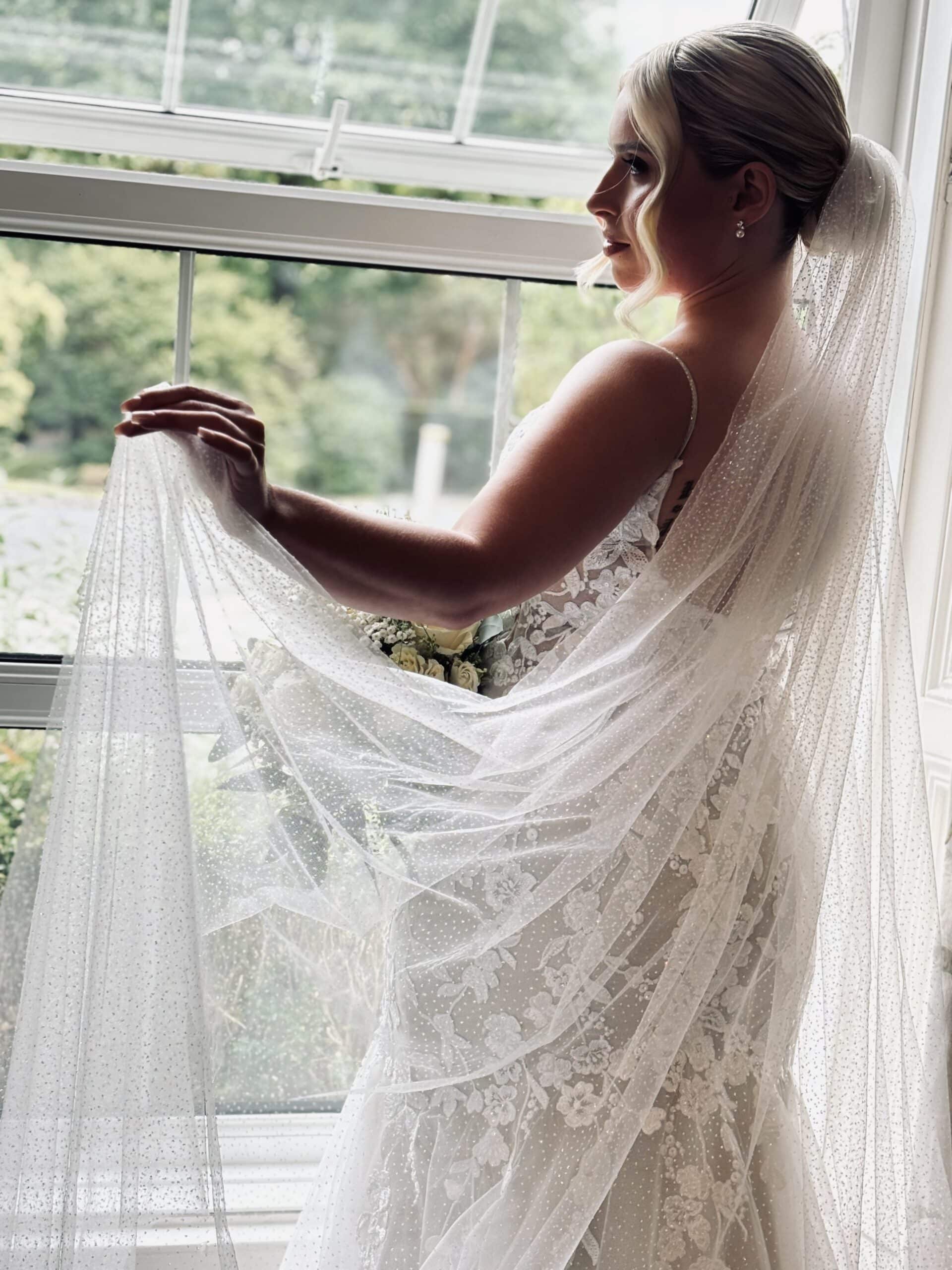 Bride in lace gown and veil, holding bouquet, gazing out window.