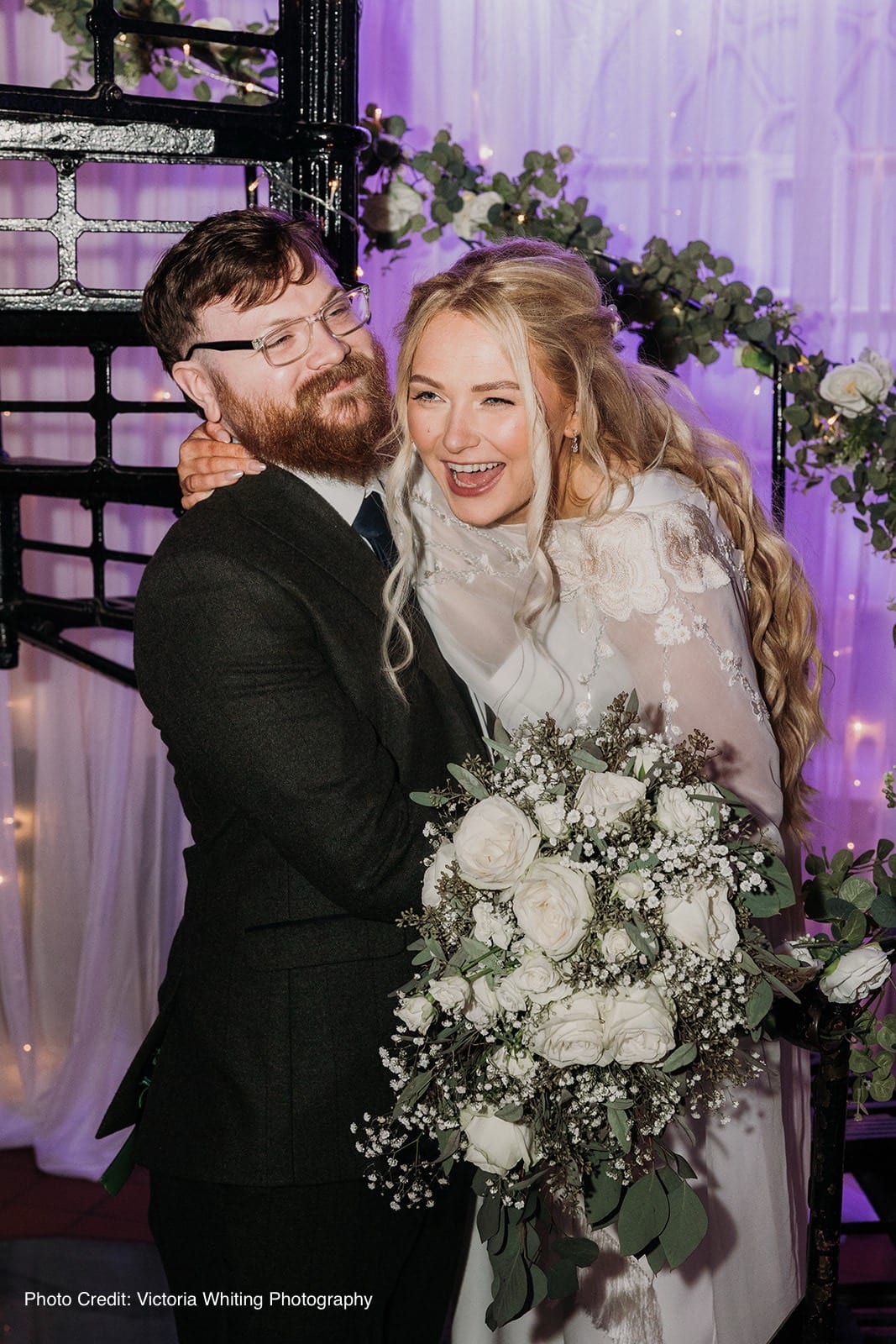 Bride and groom smiling, bride holding a bouquet with white roses, decorated arch in the background.
