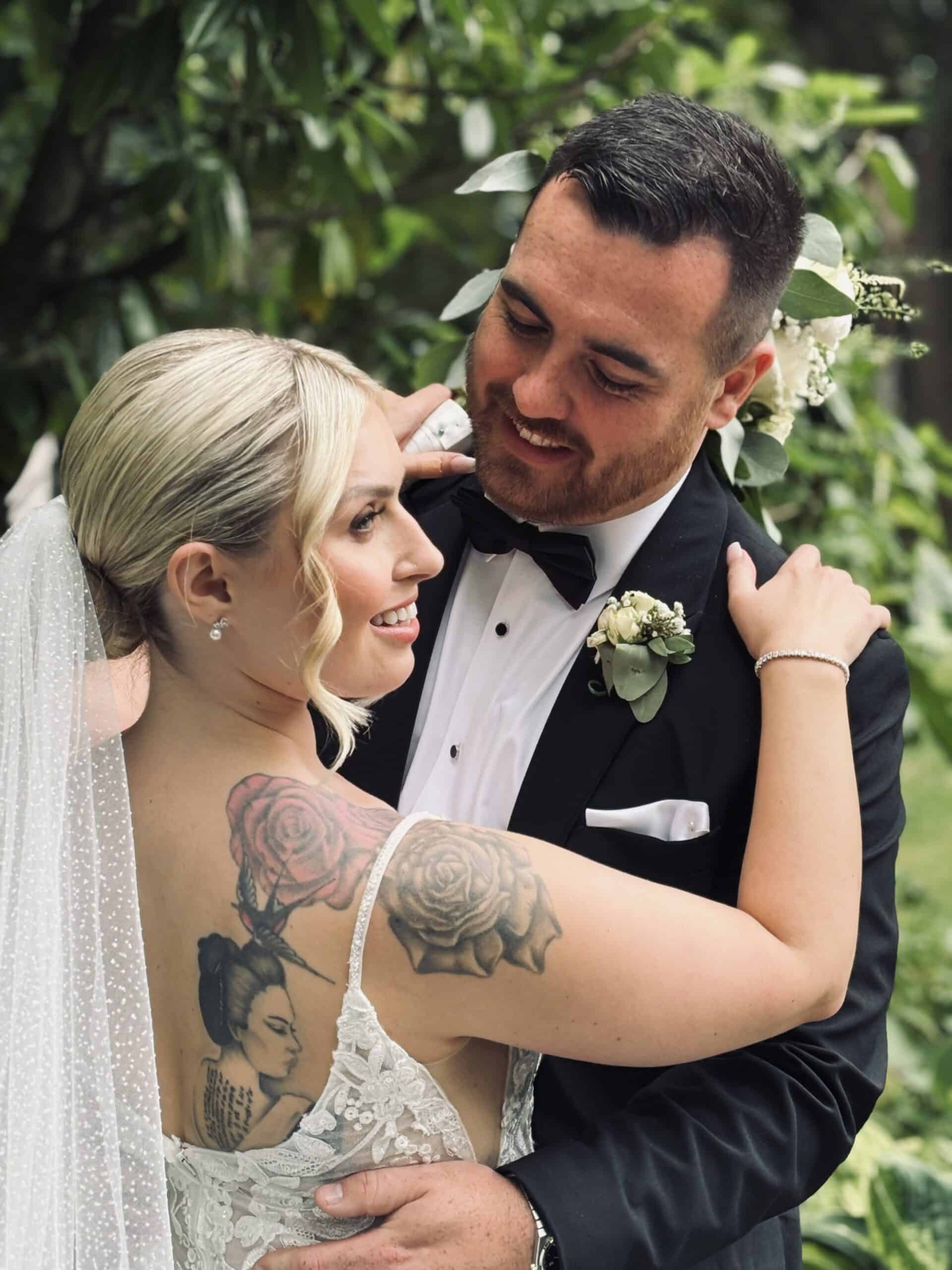Bride and groom embracing outdoors, bride showing tattoo of roses and a geisha, surrounded by greenery.