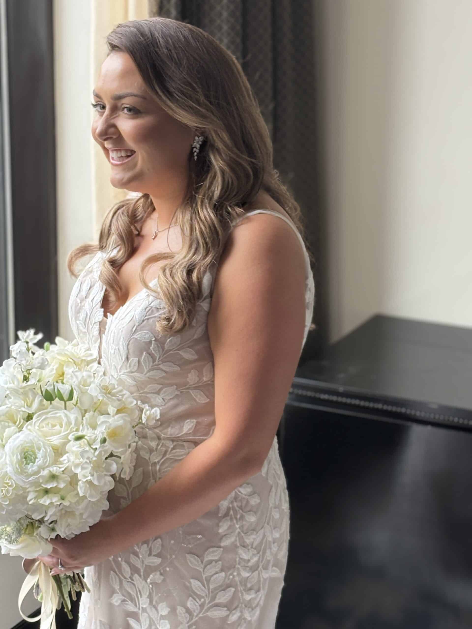 Smiling bride in an elegant white gown holding a bouquet of white flowers, standing near a window.
