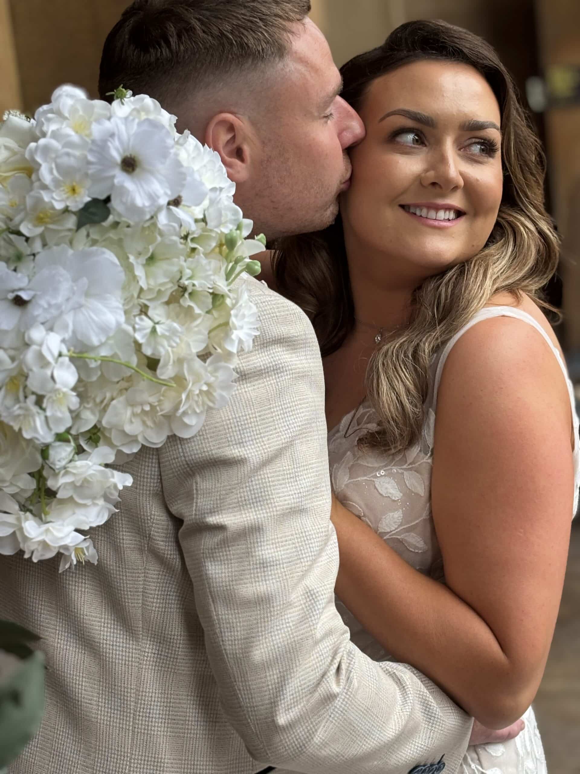 Bride and groom embracing with a bouquet of white flowers, smiling outdoors.