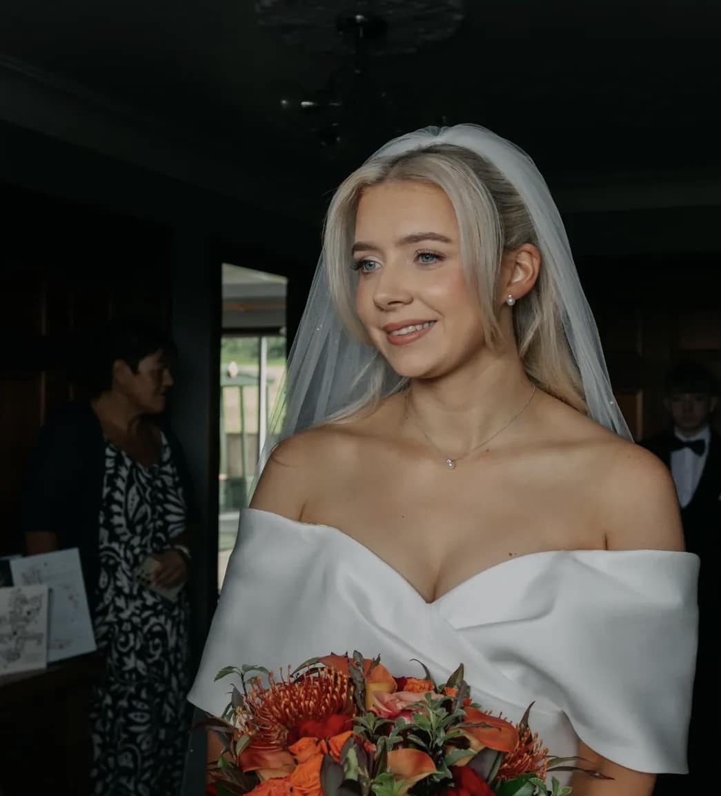 Bride in a white dress and veil holding an orange bouquet, smiling indoors.