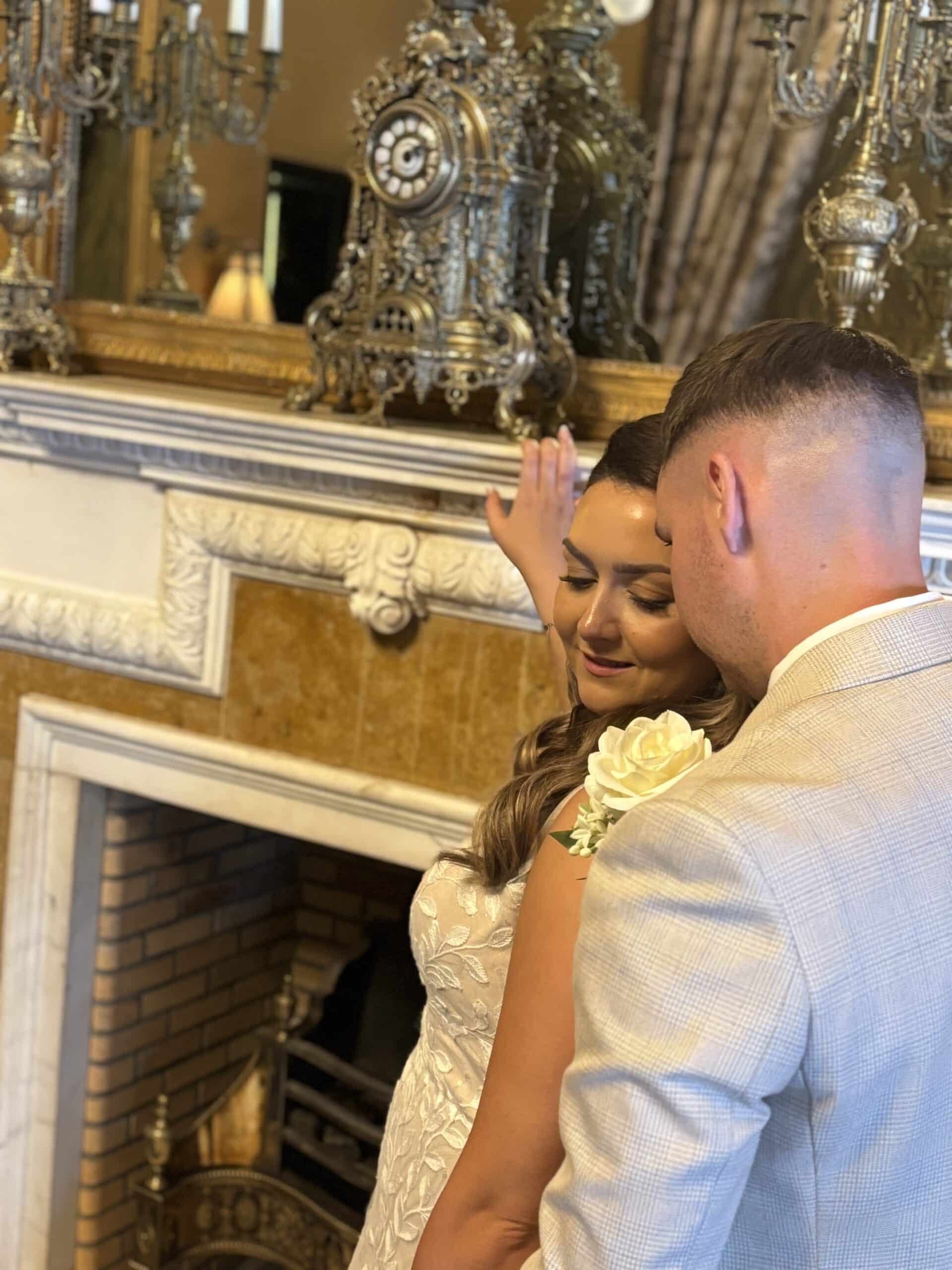 A couple in elegant attire embracing by an ornate fireplace with decorative clocks and candelabras.