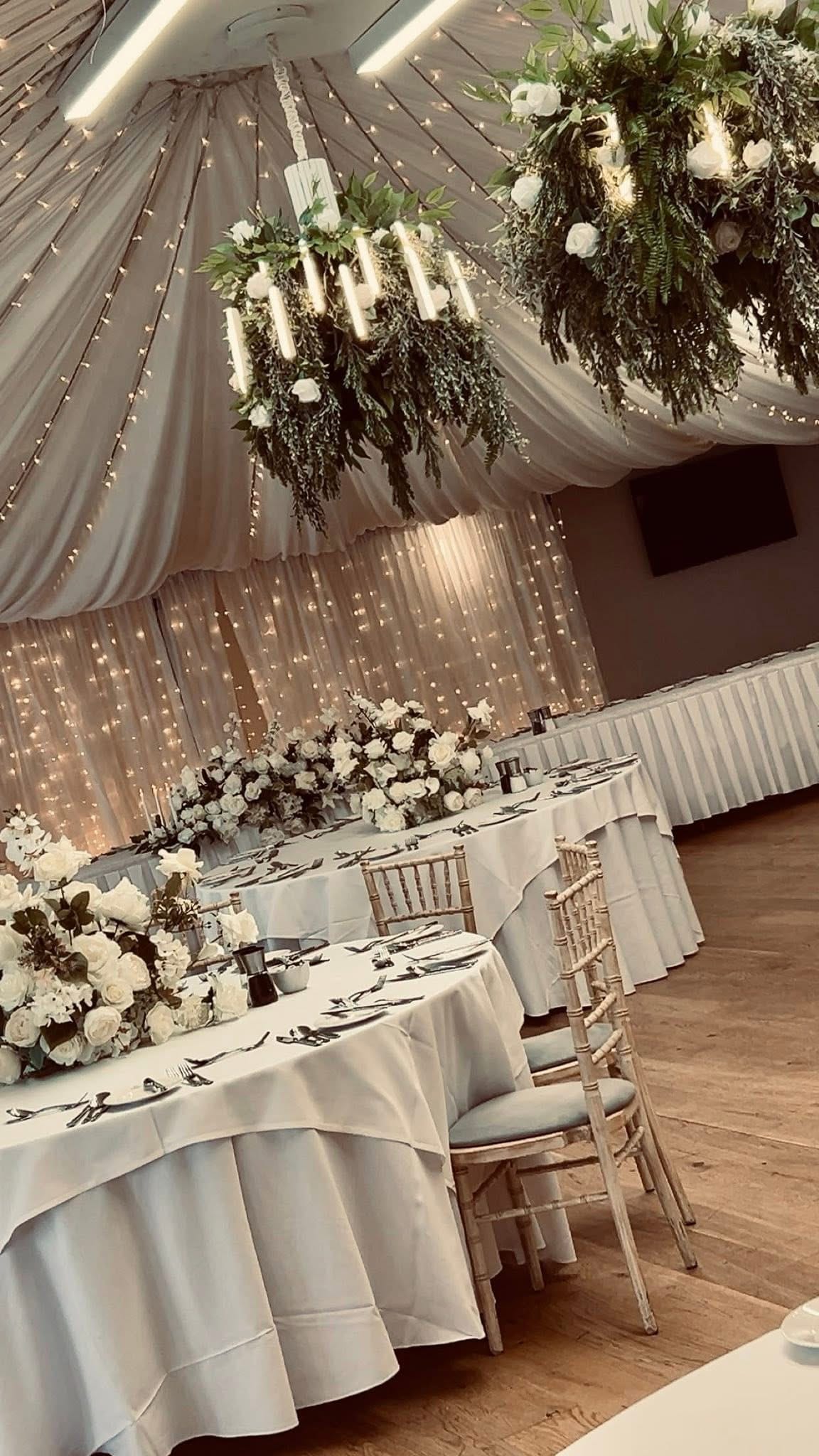 Elegantly decorated wedding venue with floral chandeliers, white draped tables, and ambient string lights.