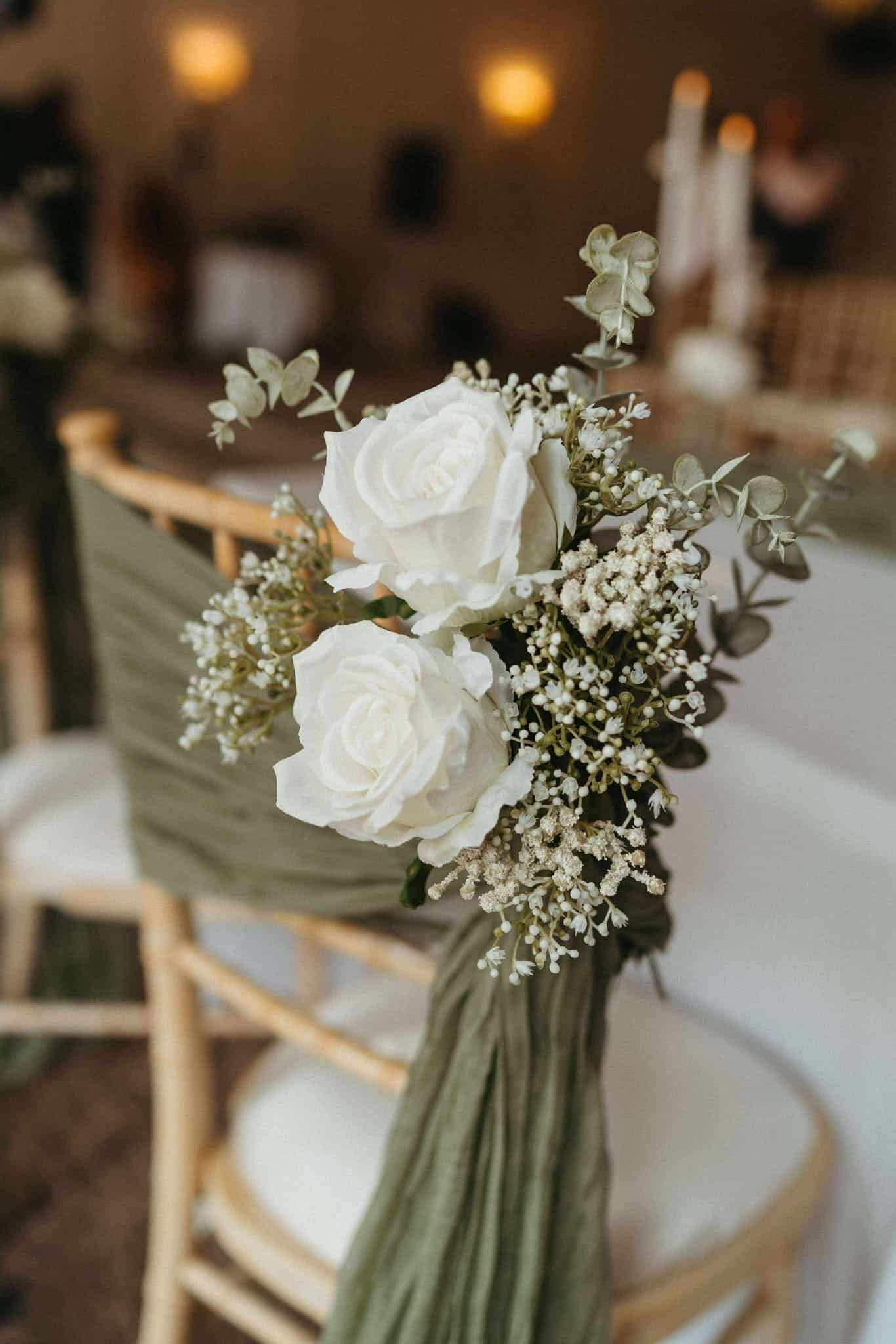 White roses and greenery tied to a wooden chair with green fabric at an event.