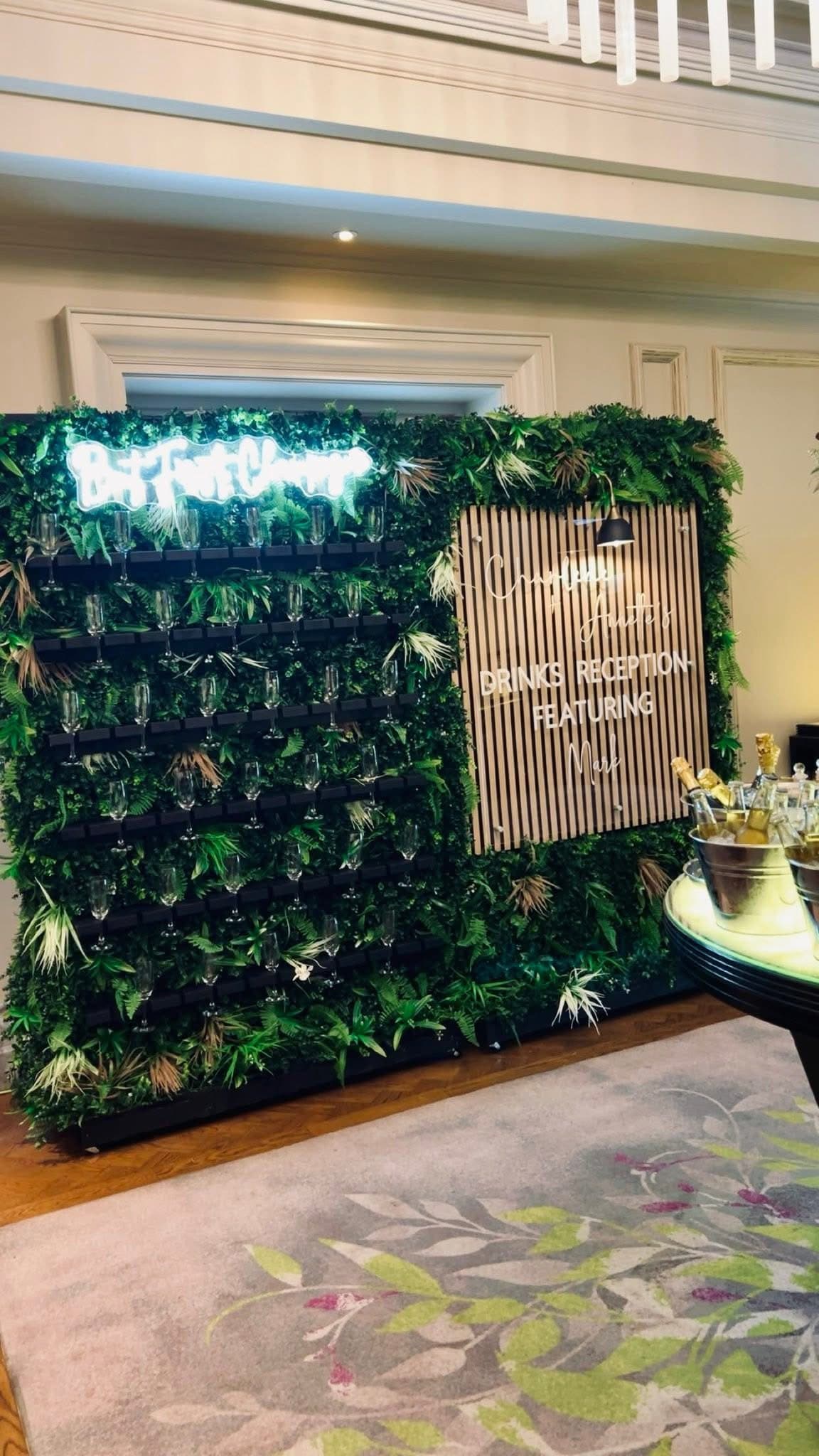Champagne wall with glasses, greenery, and neon sign at an elegant drink reception event.