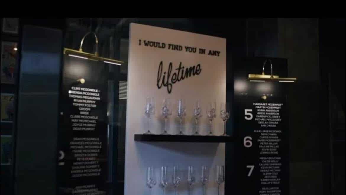 Shelf with wine glasses and sign reading "I would find you in any lifetime" illuminated by wall lights.