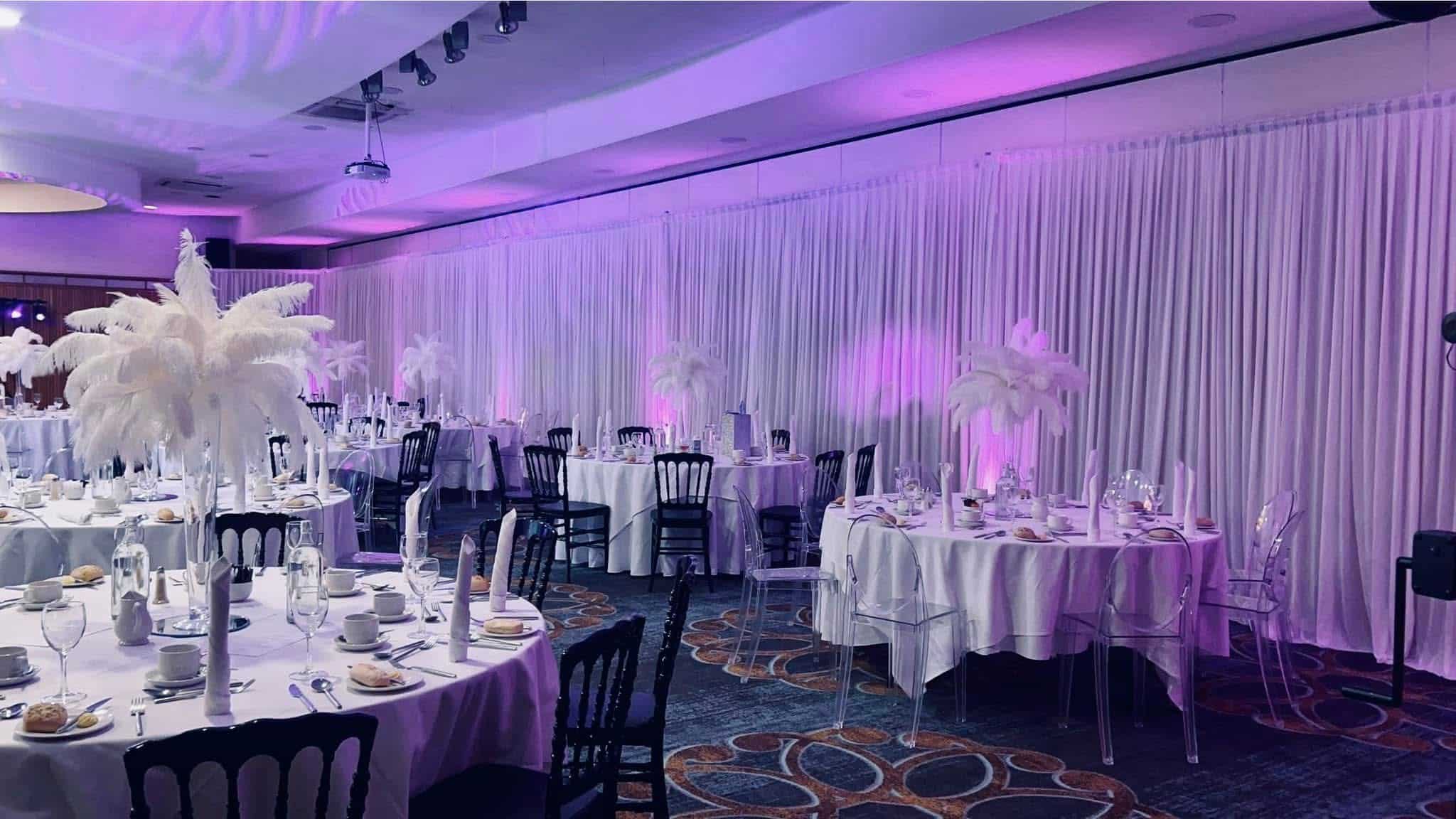 Elegant banquet hall with round tables, white tablecloths, feather centerpieces, and purple ambient lighting.