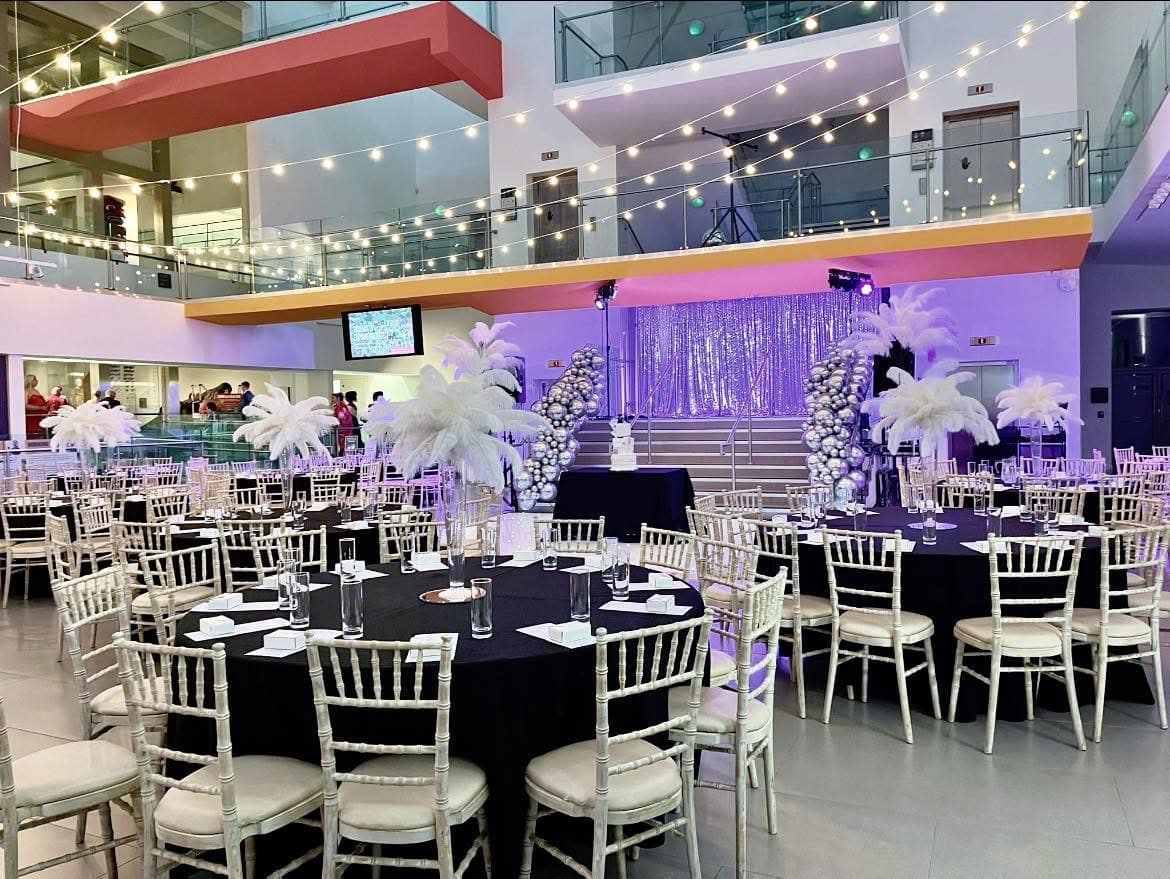 Event space with round tables, white chairs, feather centerpieces, a stage with silver backdrop, and string lights overhead.