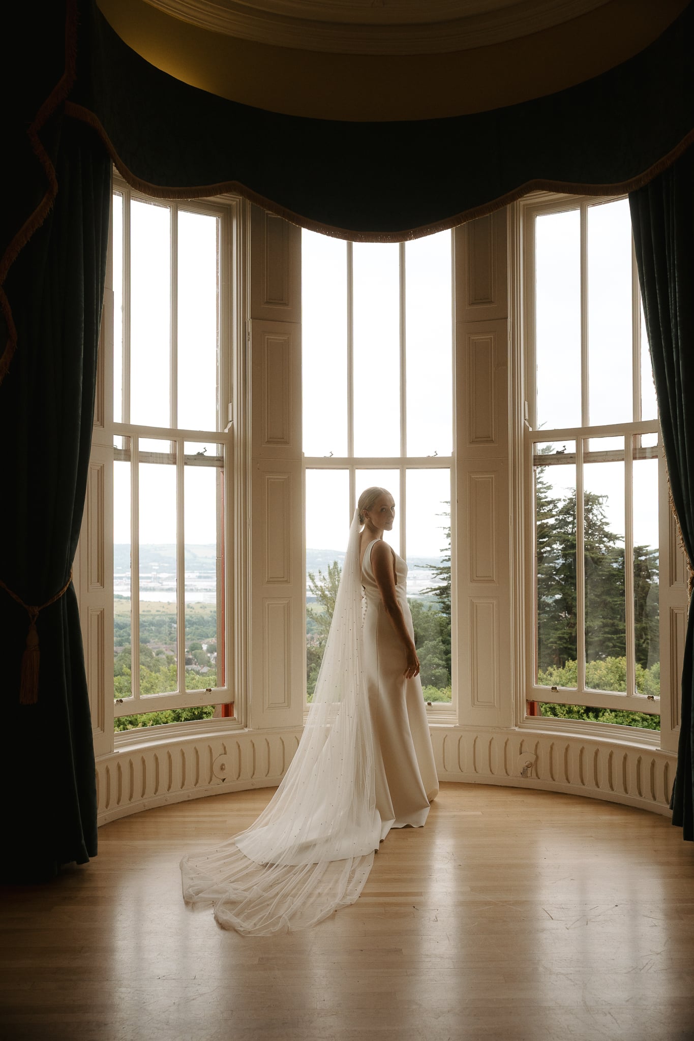 Bride in elegant gown and veil stands near large windows overlooking a scenic view.
