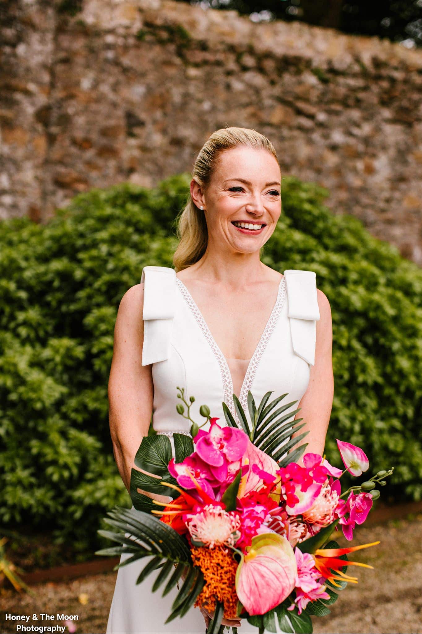 Smiling bride holding a vibrant tropical bouquet with pink and orange flowers against a stone wall and greenery background.