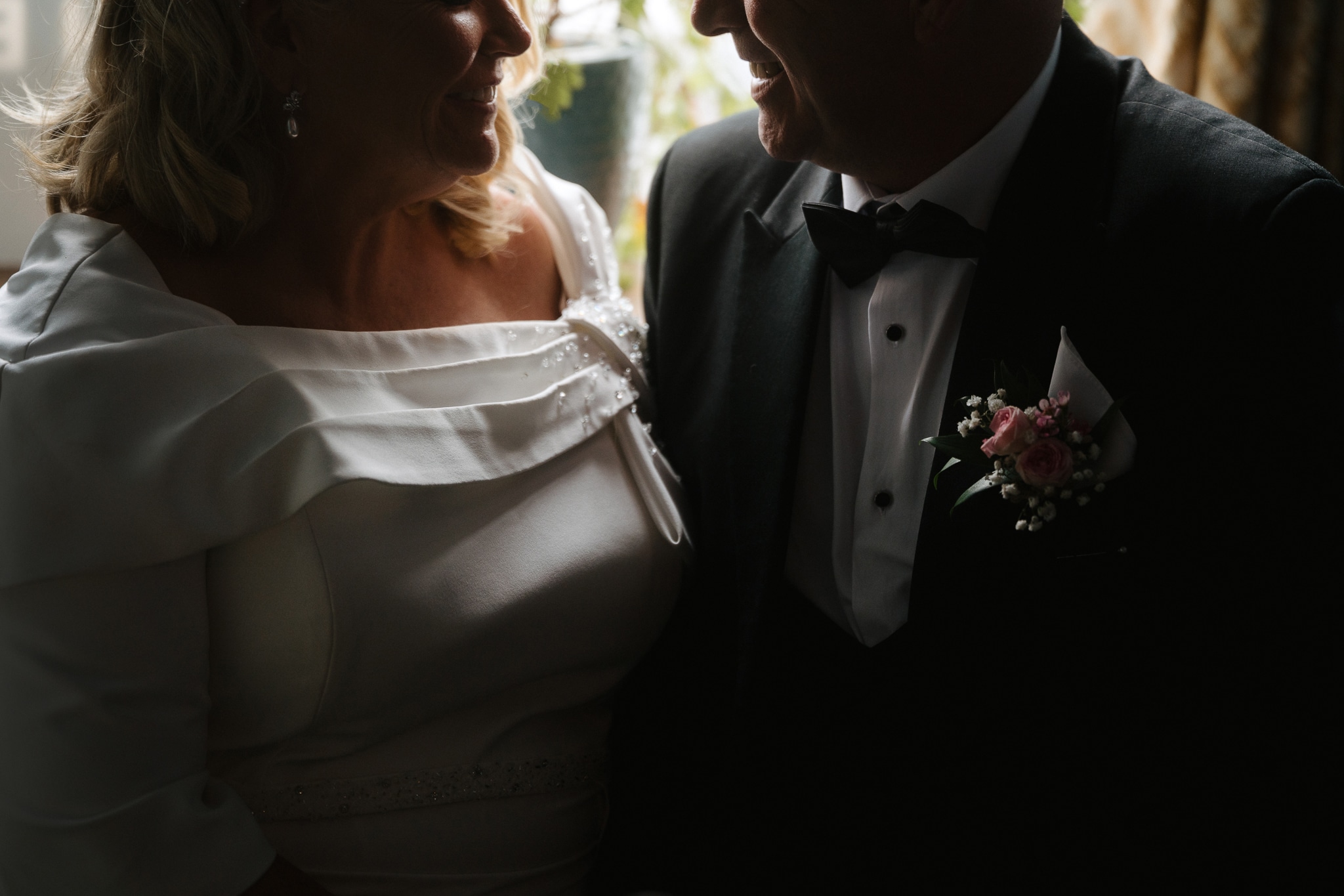Bride and groom smiling at each other, wearing wedding attire with floral boutonniere visible.