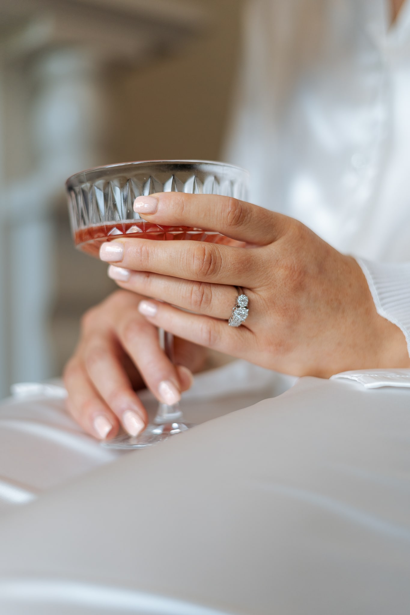 Close-up of a hand holding a crystal glass with a ring on the finger, wearing a silky white garment.