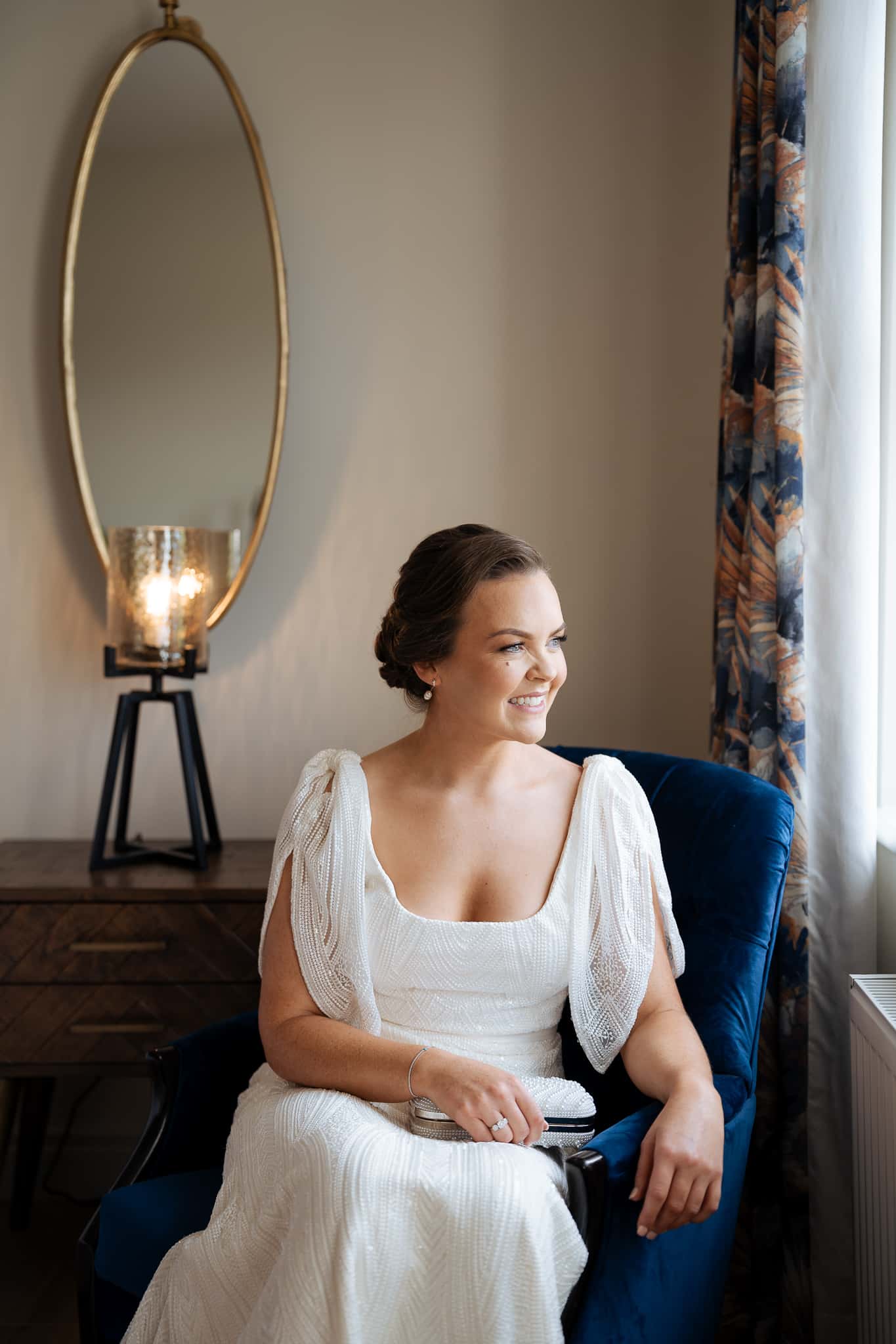Bride in elegant white dress sitting, smiling, and holding a clutch, with a mirror and lamp in the background.