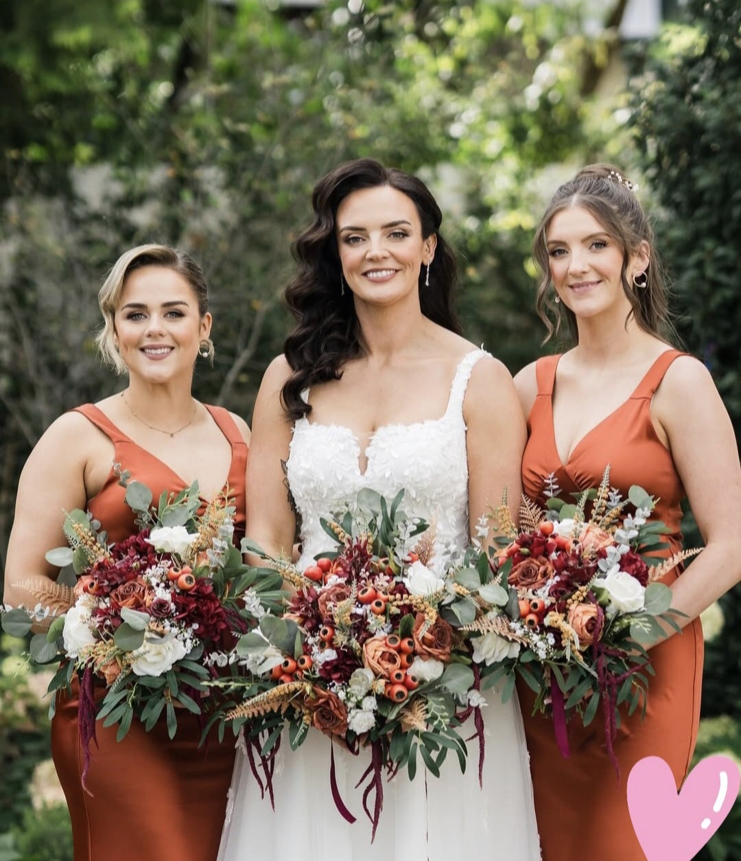 Bride with two bridesmaids holding floral bouquets in an outdoor setting.