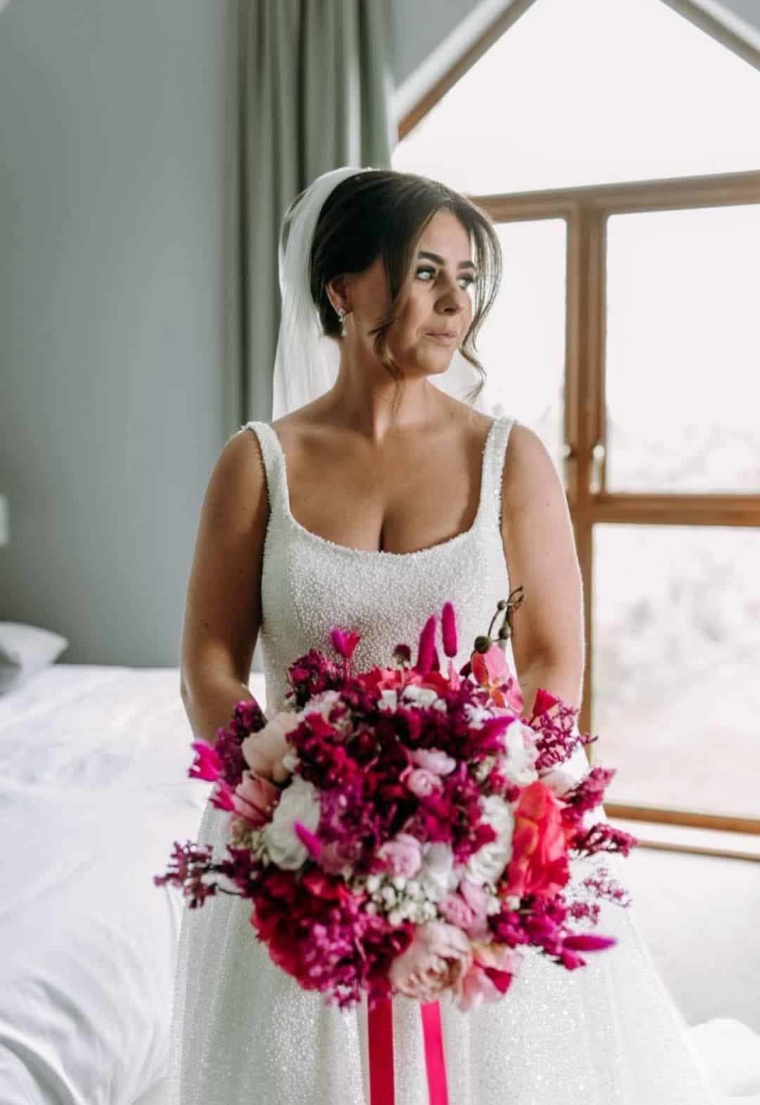 Bride in a white dress holding a vibrant bouquet with pink and red flowers, standing by a window.