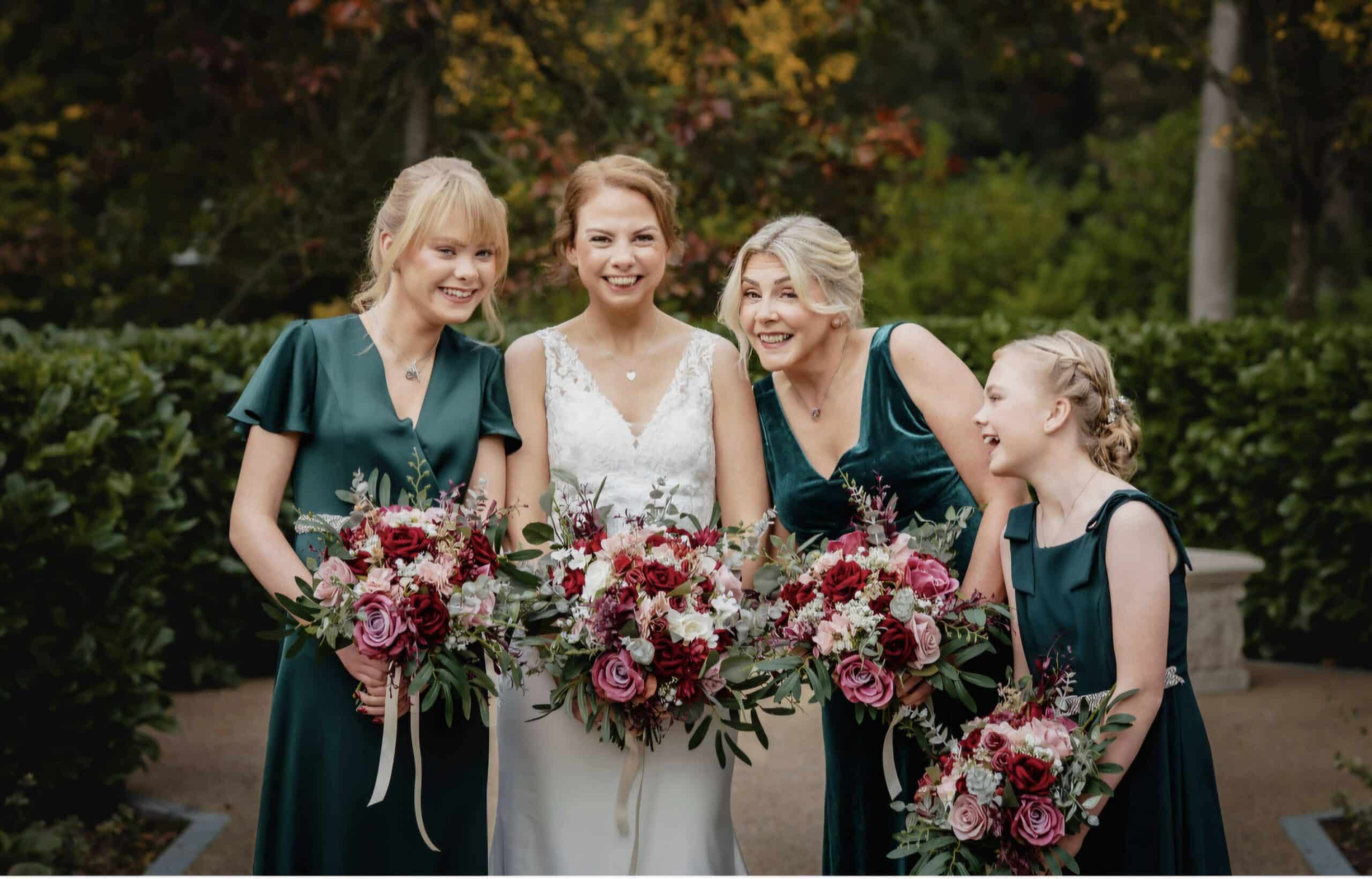 Bride and bridesmaids in green dresses holding floral bouquets, smiling outdoors.