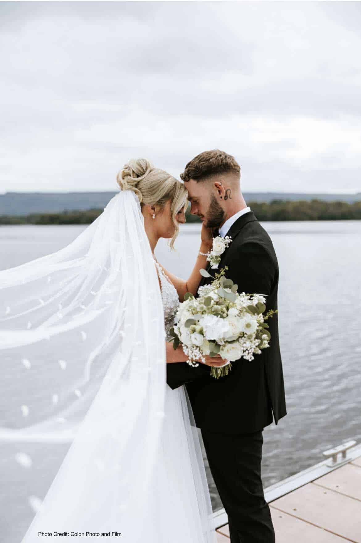 Bride and groom embrace on a lakeside dock, bride holding bouquet and veil flowing.
