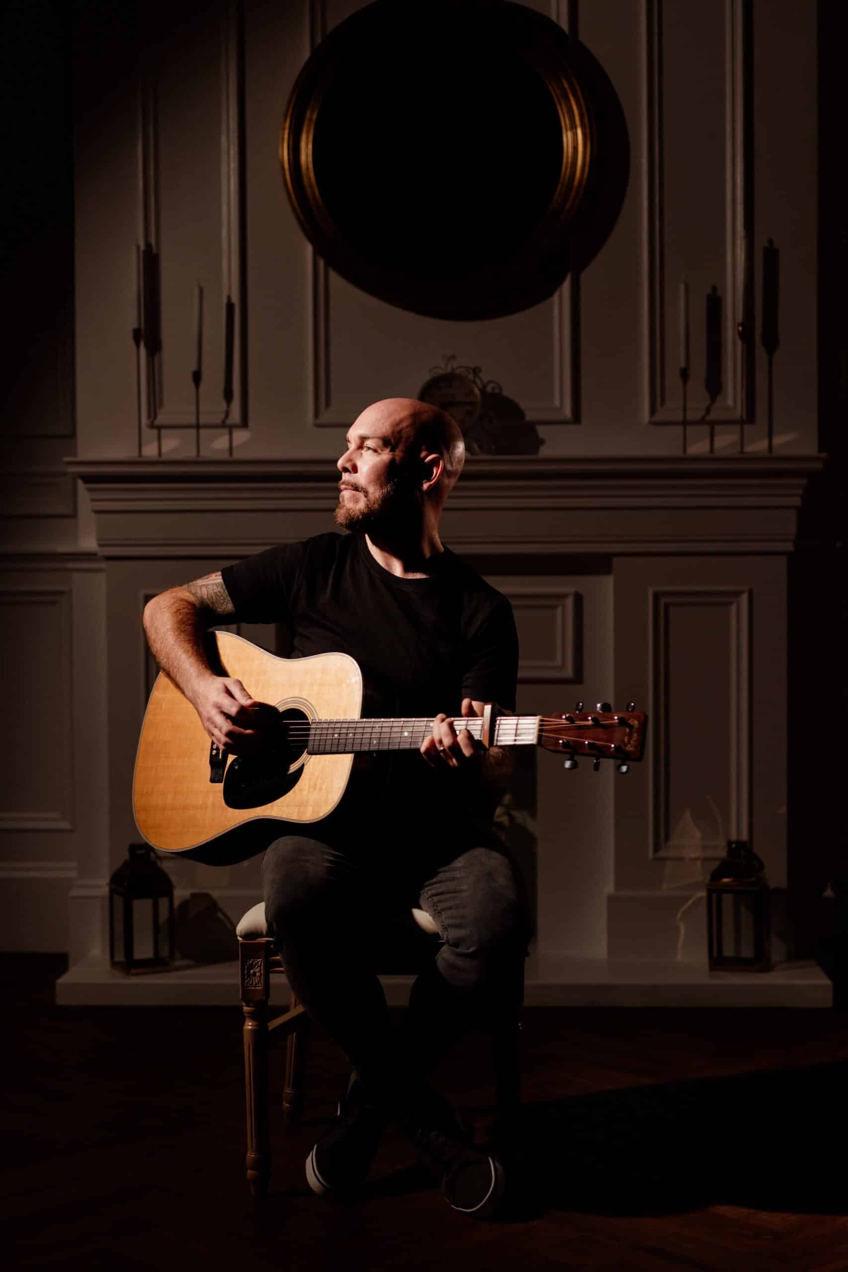 Bald man playing acoustic guitar in dimly lit room with candles and ornate wall.