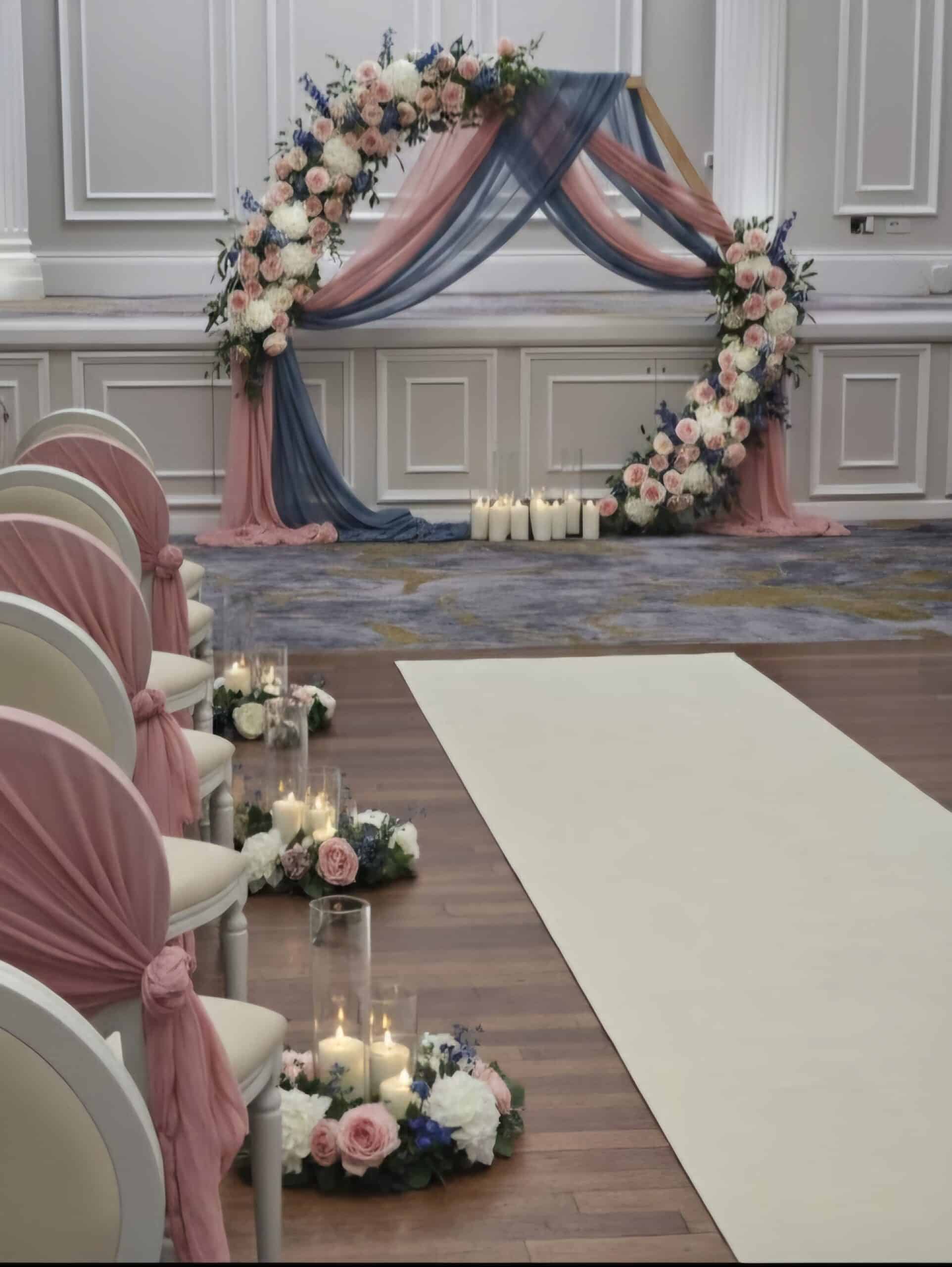 Elegant wedding aisle with floral arch, candles, and pink draped chairs in a beautifully decorated room.