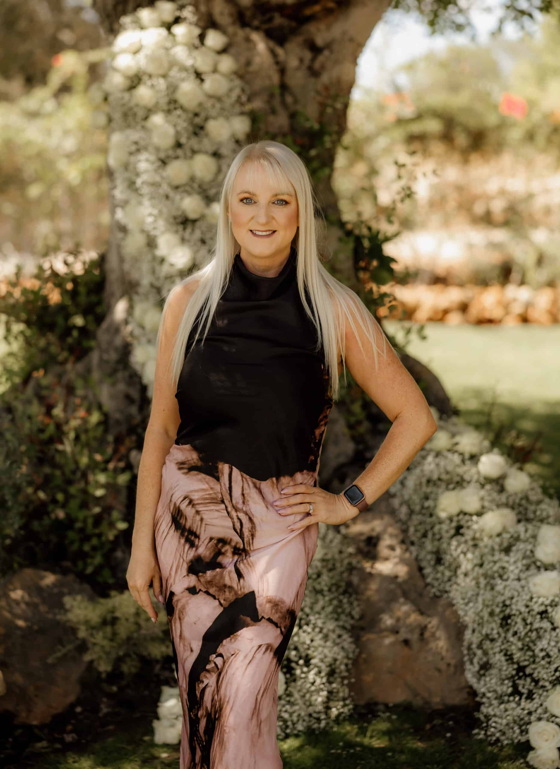 Woman in a stylish dress poses outdoors near a tree adorned with white flowers.