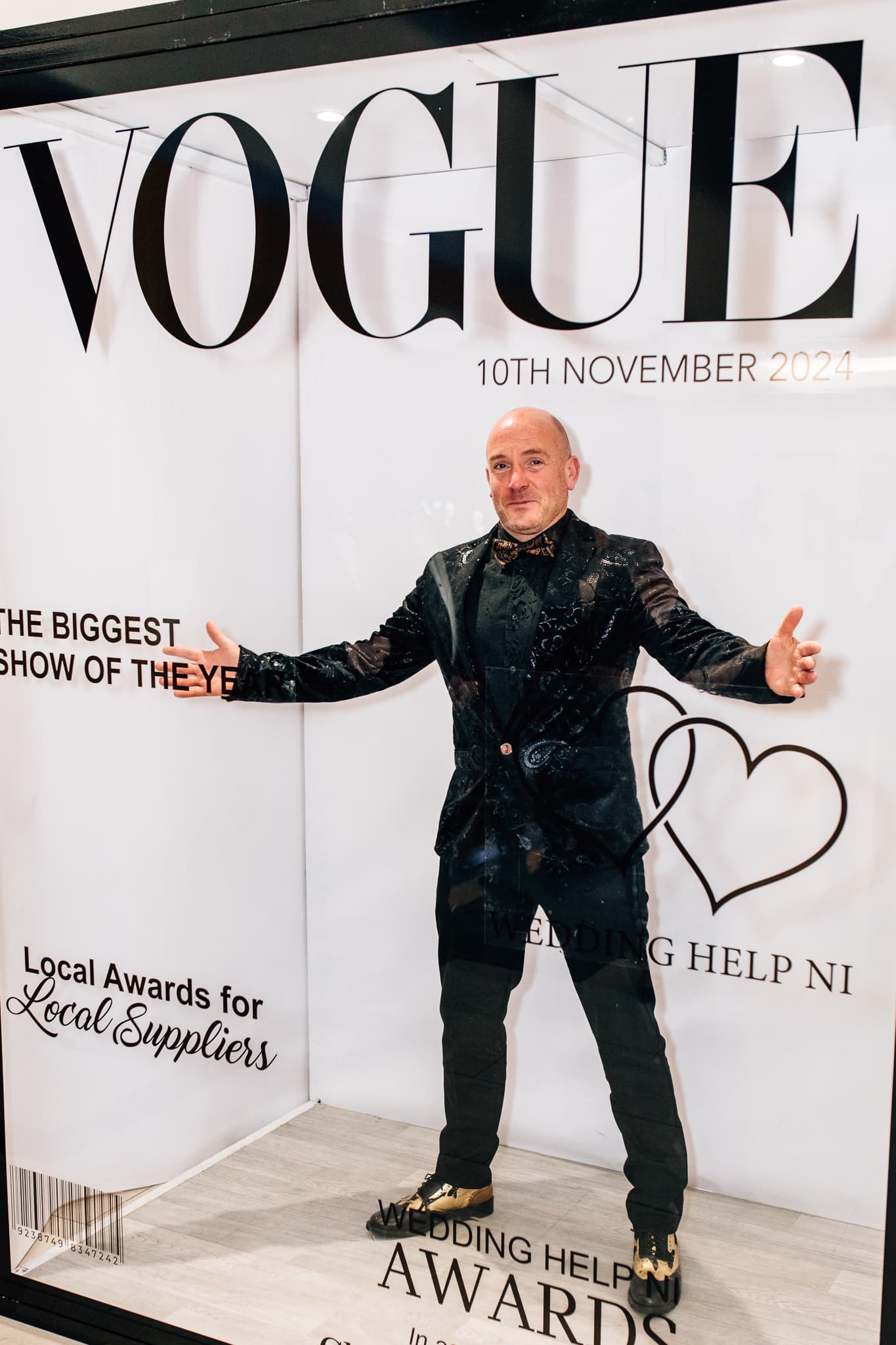 Man in formal attire posing at Wedding Help NI Awards display with Vogue branding.