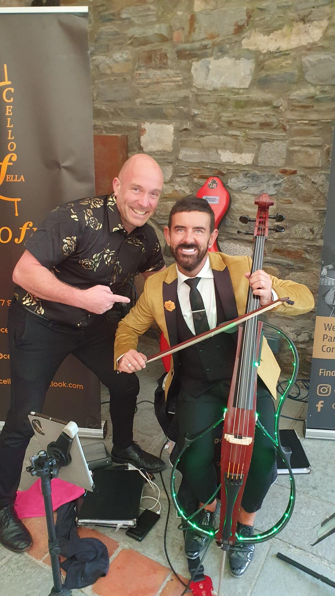 Two men smiling; one is holding a cello. Stone wall backdrop with promotional banners.