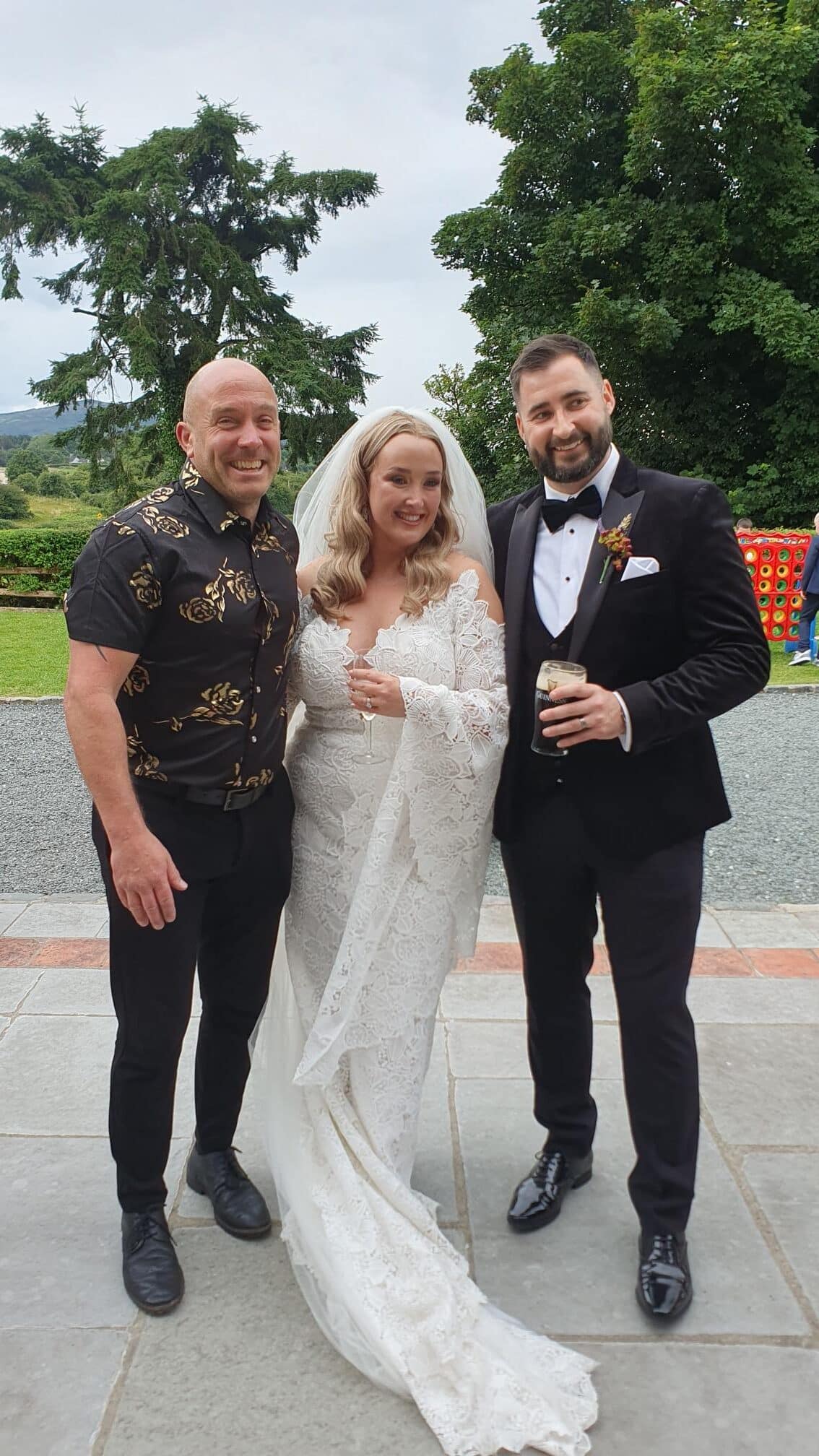 Bride and groom pose with a guest at an outdoor wedding, greenery in the background.