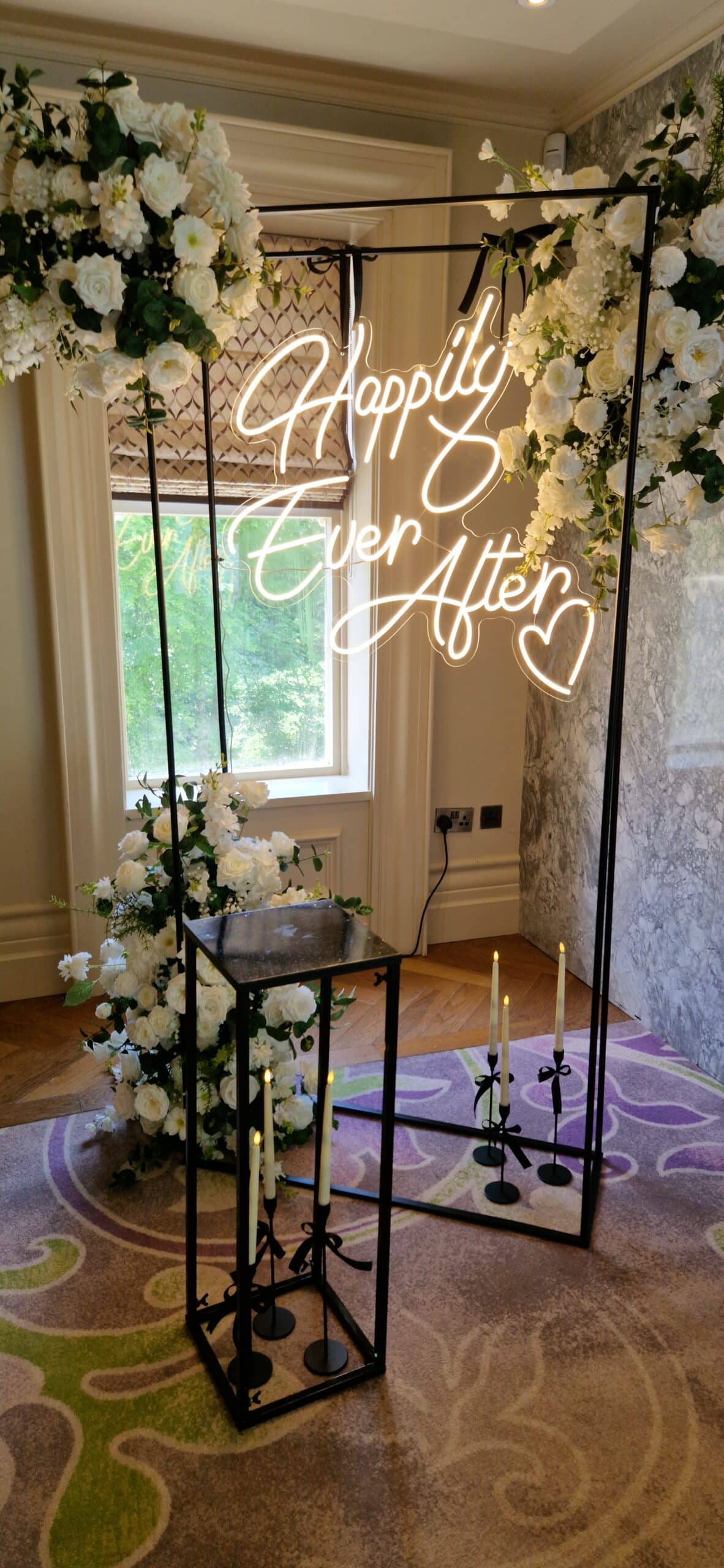 Neon "Happily Ever After" sign surrounded by white floral arrangements and candles in a decorative setting.