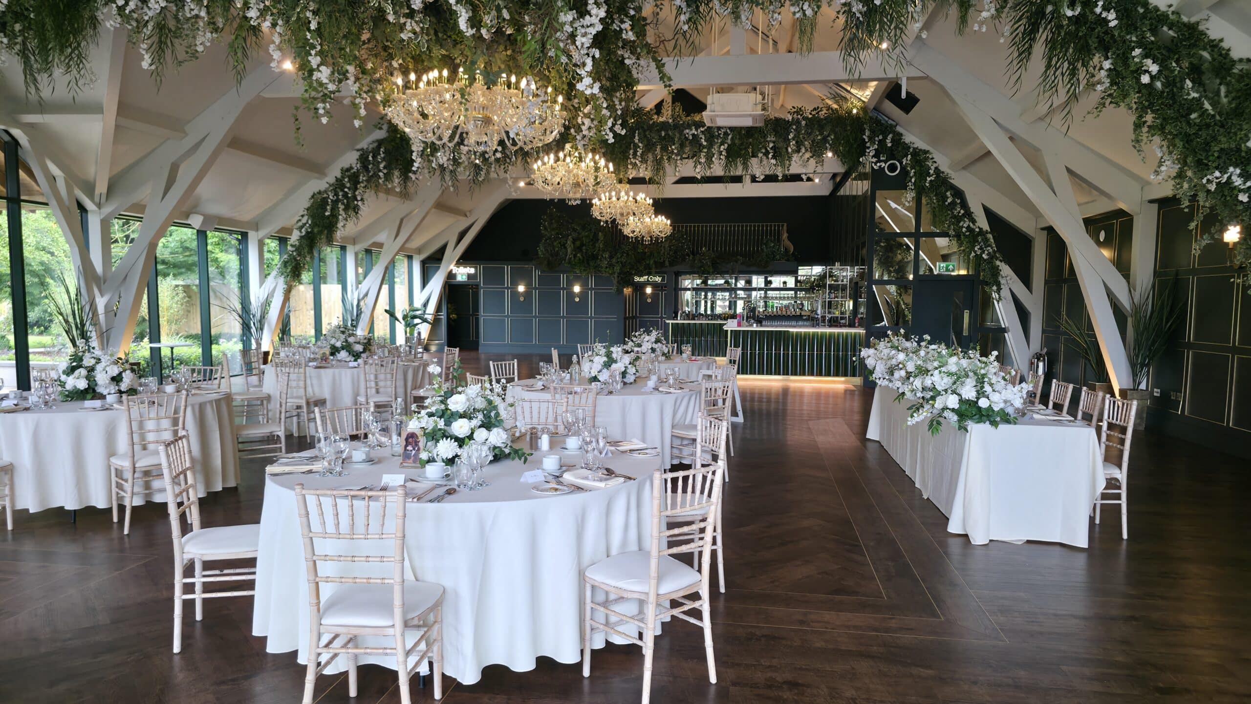 Elegant wedding reception hall with flower arrangements, chandeliers, round tables, and chairs under a decorated ceiling.