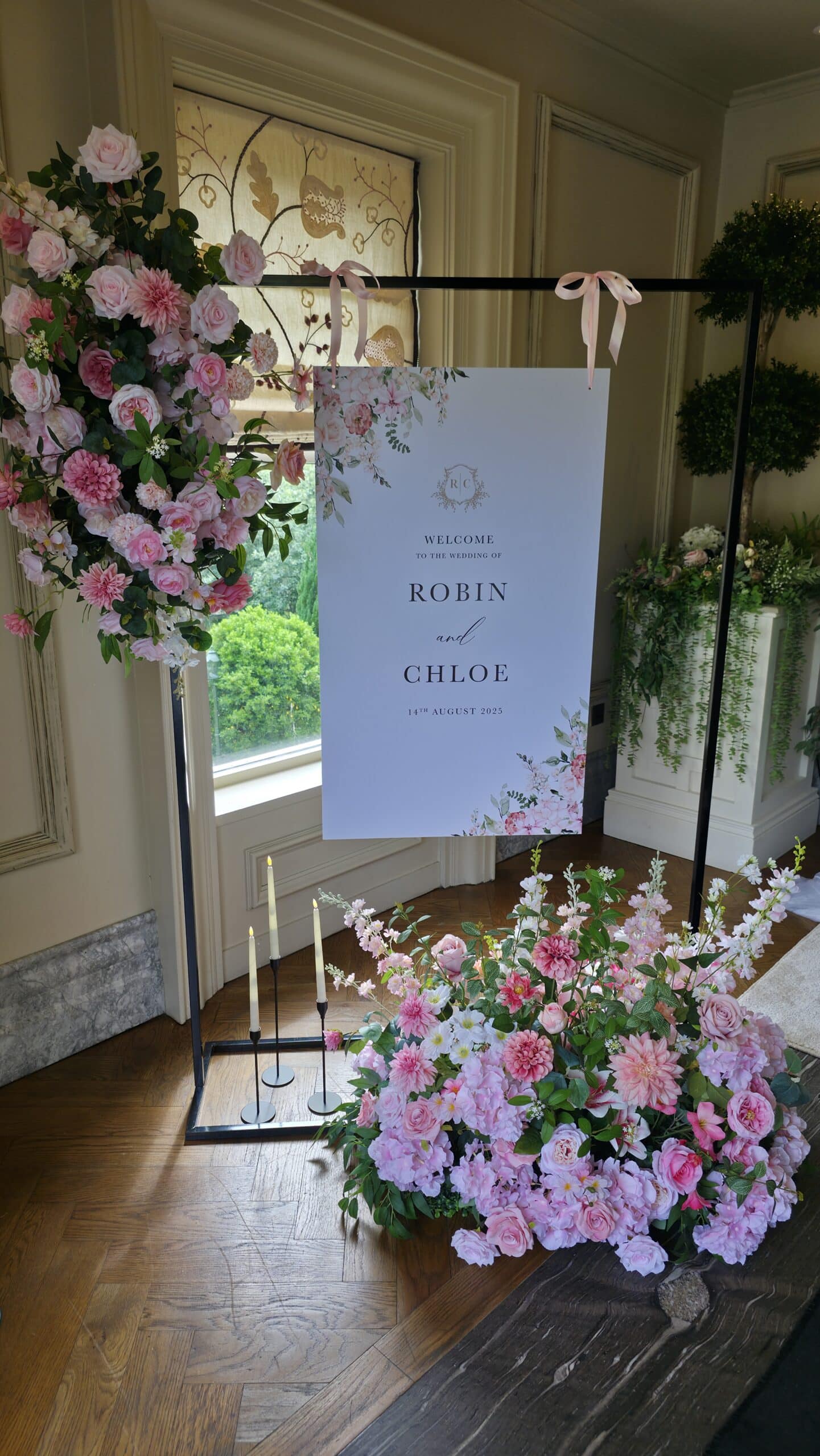 Wedding welcome sign for Robin and Chloe on 14th August 2023, surrounded by pink floral arrangements indoors.
