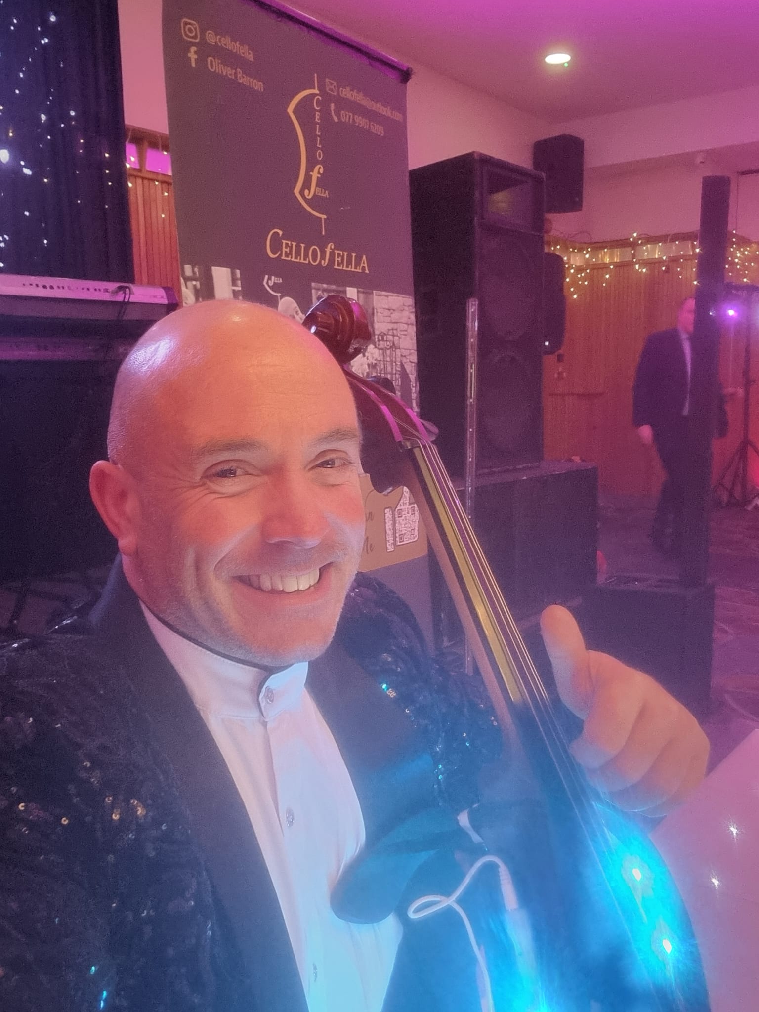 Man smiling with cello, wearing sequin jacket; "Cello Fella" banner in background at event.