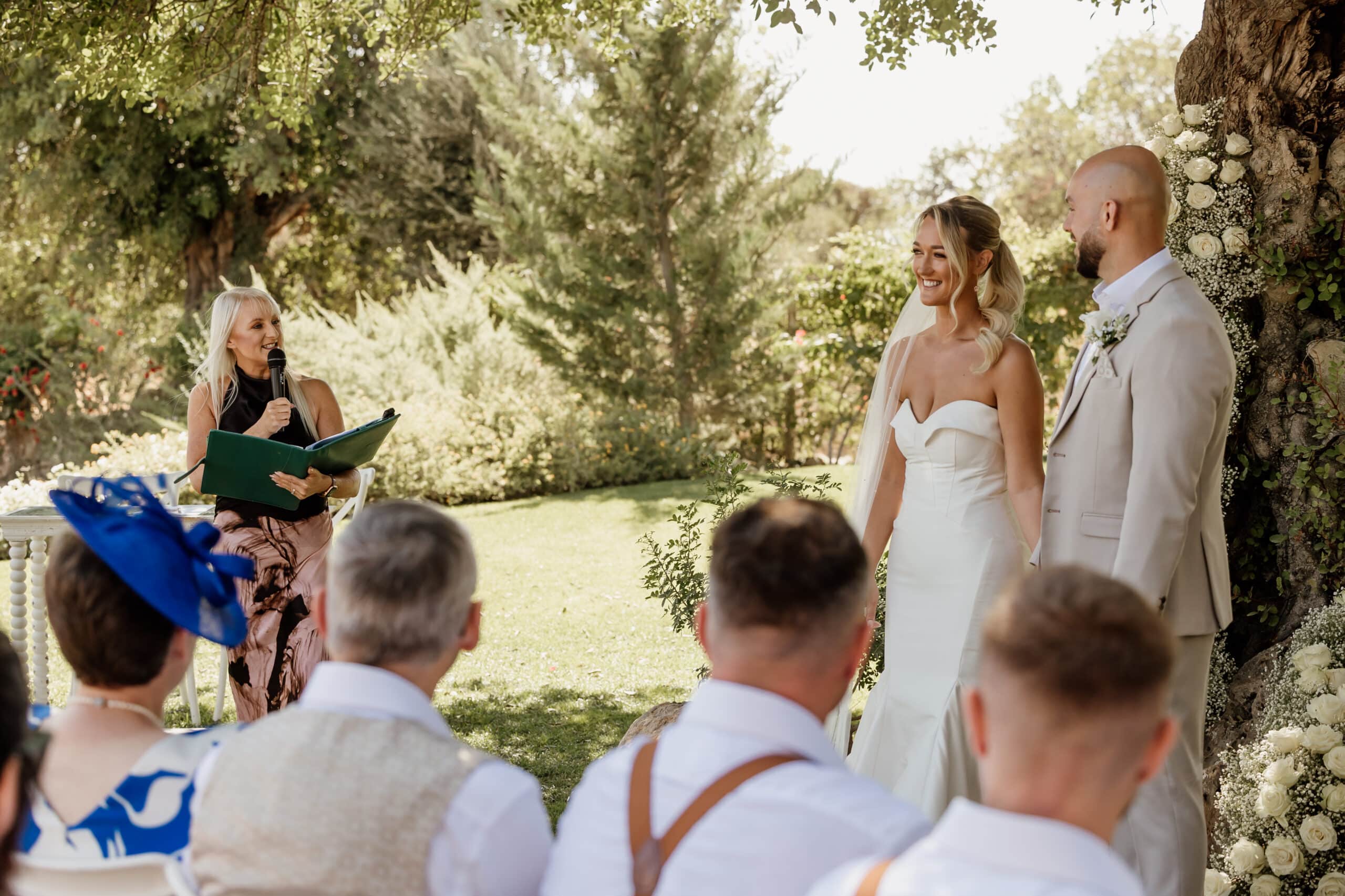 Bride and groom smiling during outdoor wedding ceremony under tree, officiant speaking.