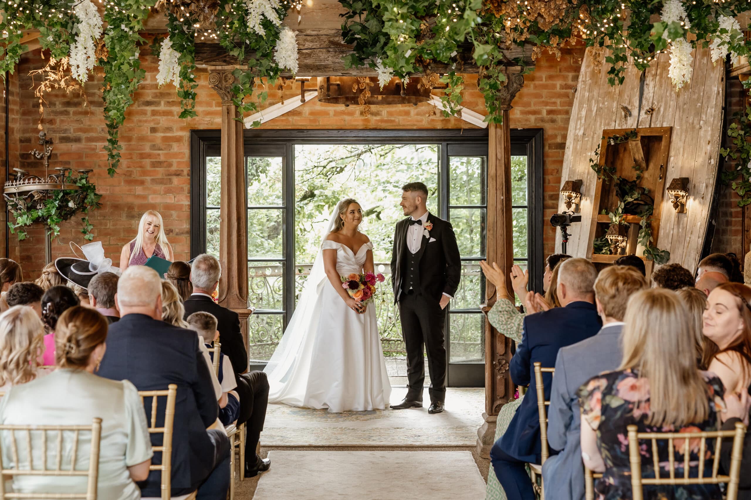 Bride and groom standing at wedding ceremony with guests seated, in a decorated rustic venue.