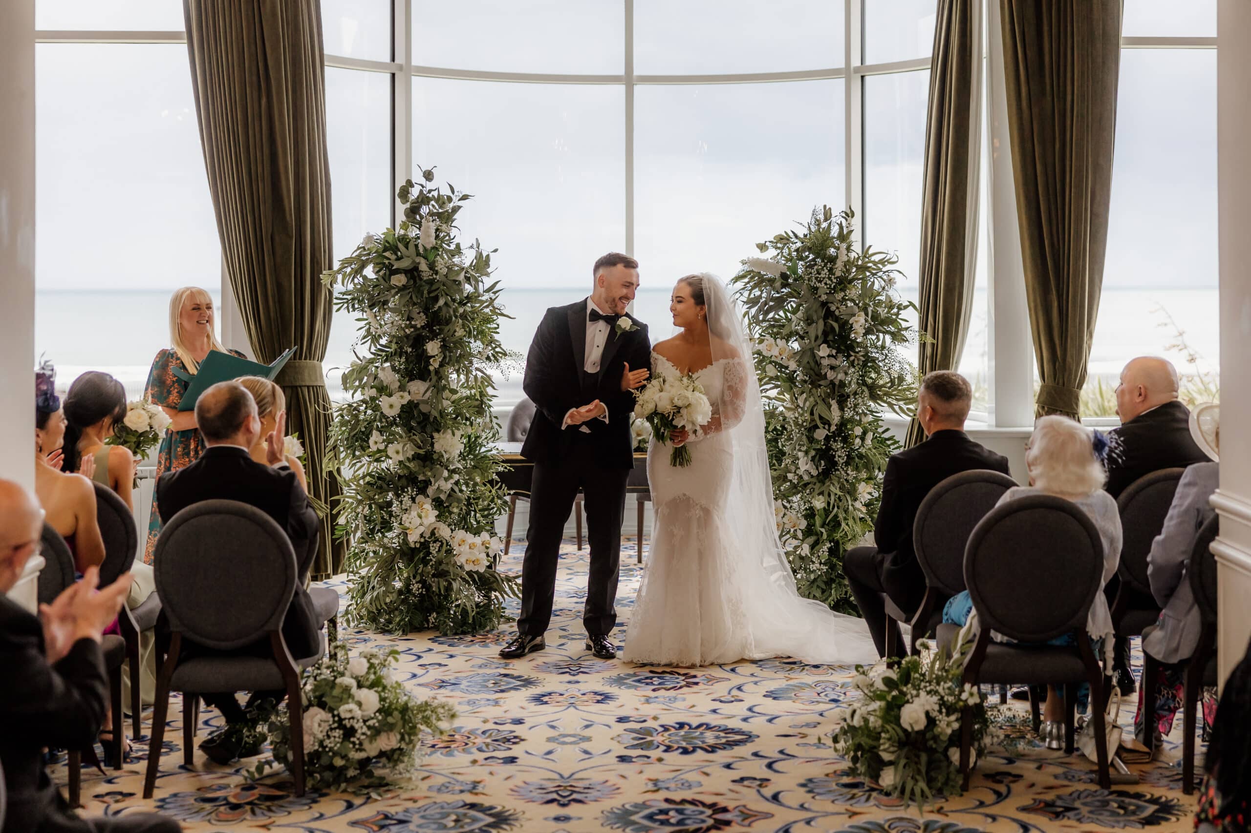Bride and groom standing at an indoor wedding ceremony, surrounded by guests and floral decorations.