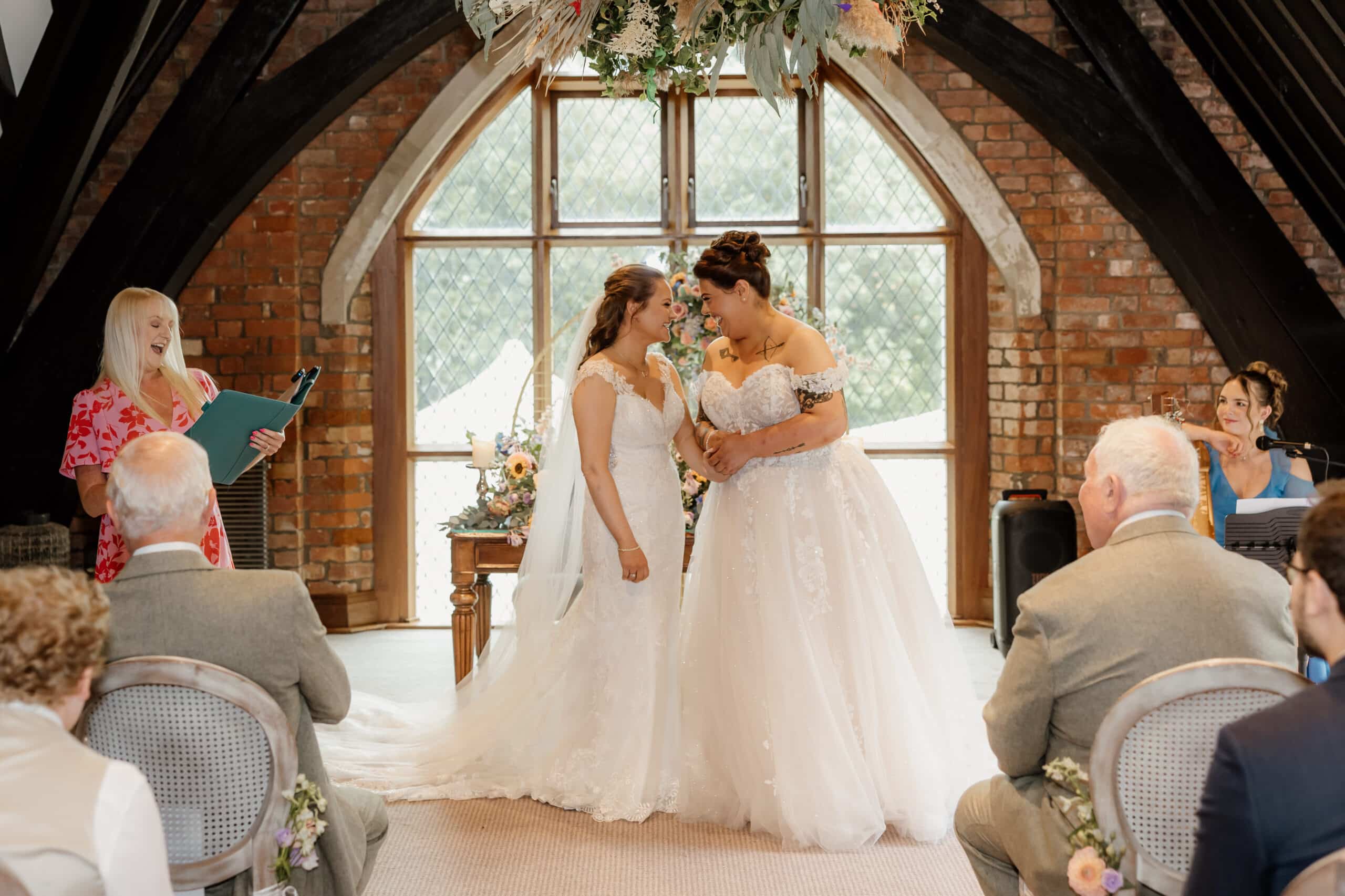 Two brides holding hands during a wedding ceremony, with guests seated nearby.