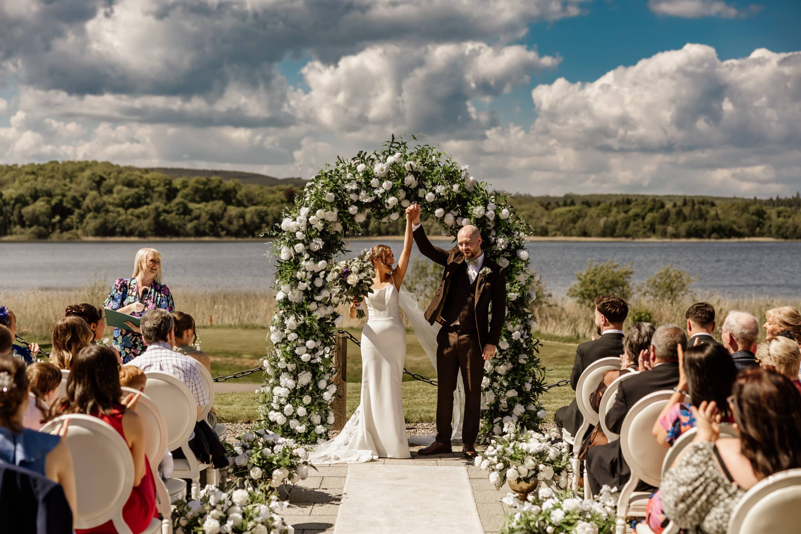 Bride and groom celebrate under a floral arch during an outdoor wedding ceremony by a lake, with guests seated nearby.