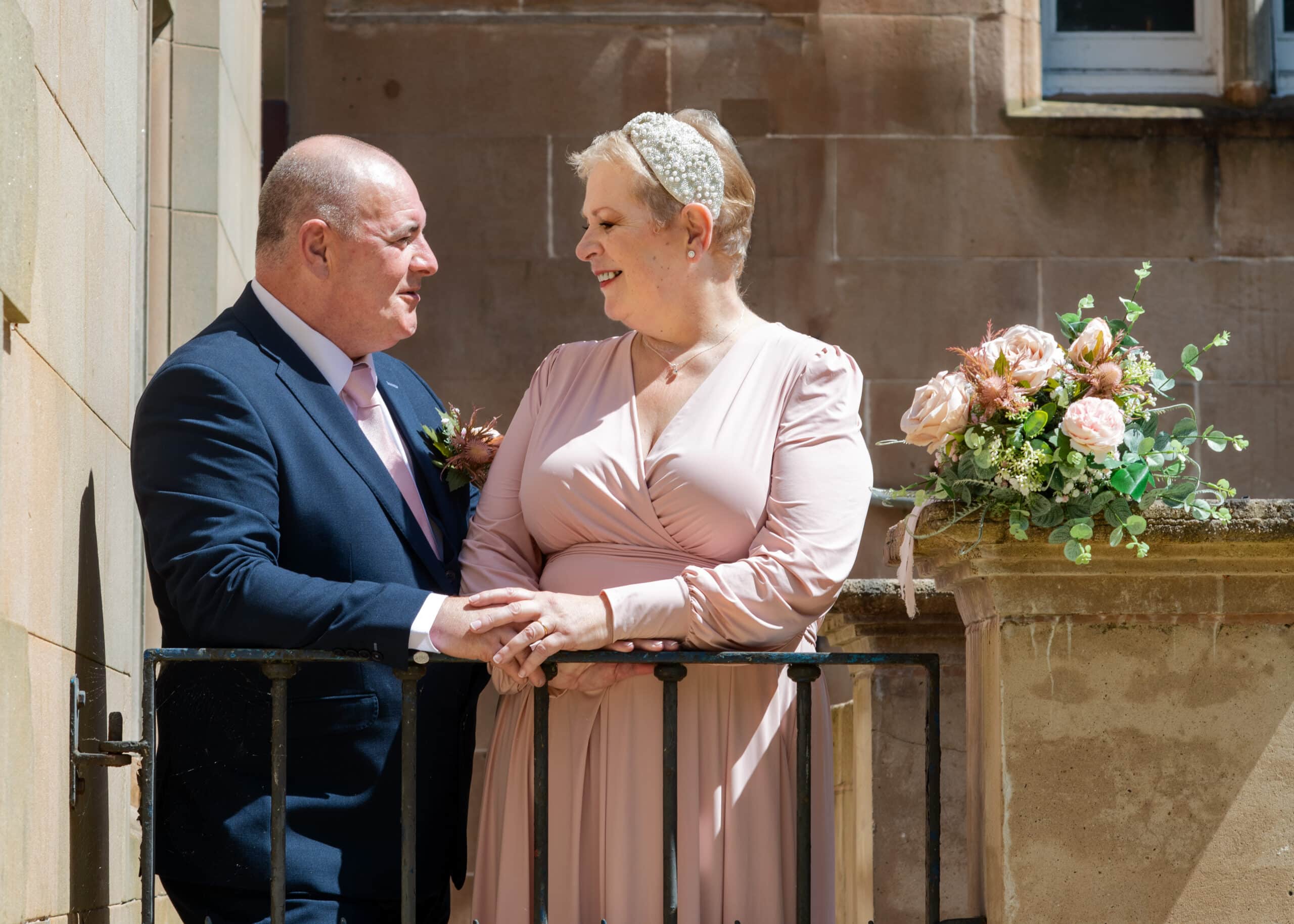 Couple smiling at each other on balcony, woman in pink dress, man in suit, with bouquet of flowers nearby.