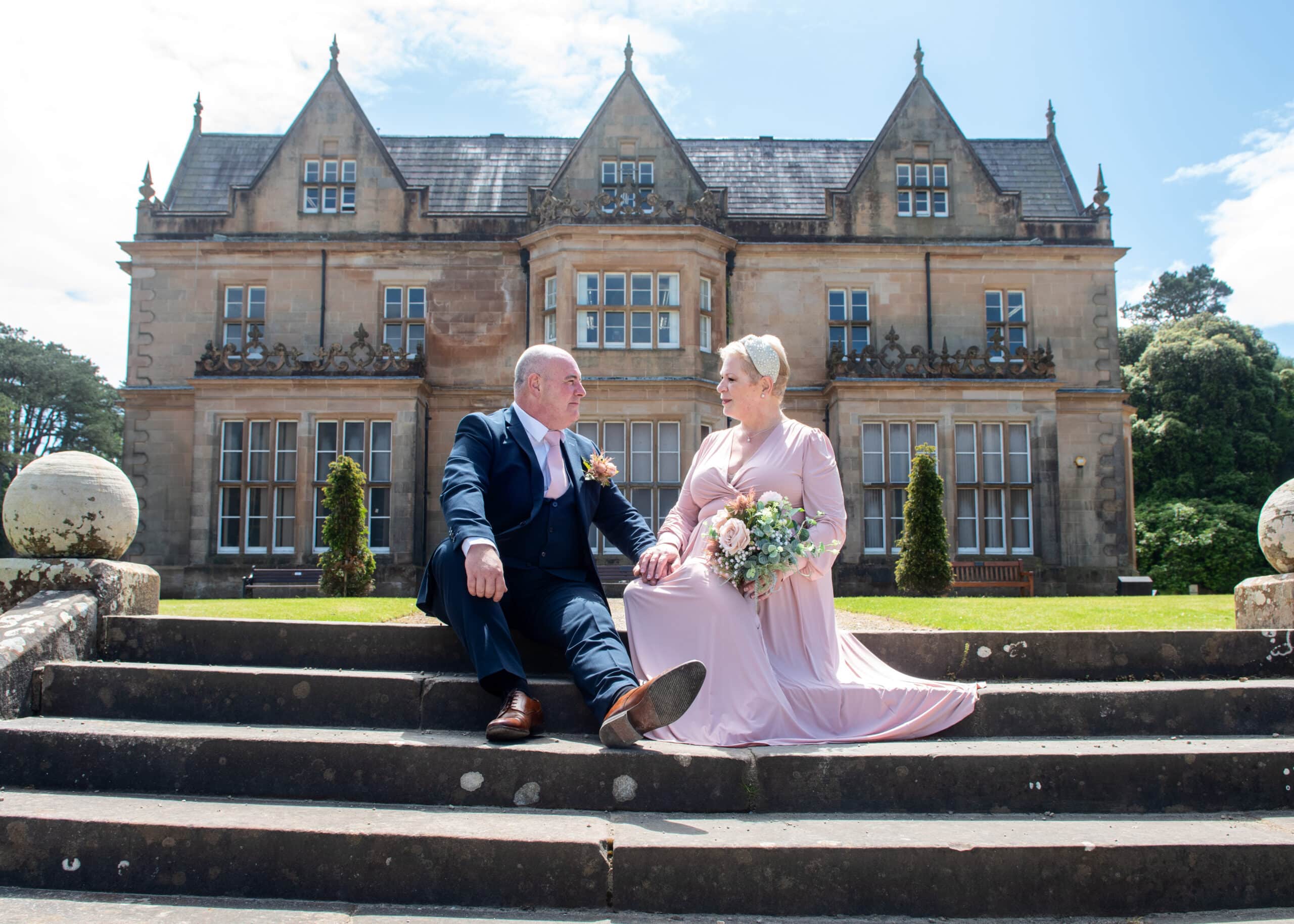 Couple sitting on steps in front of a historic mansion, dressed in formal attire, holding hands and a bouquet.