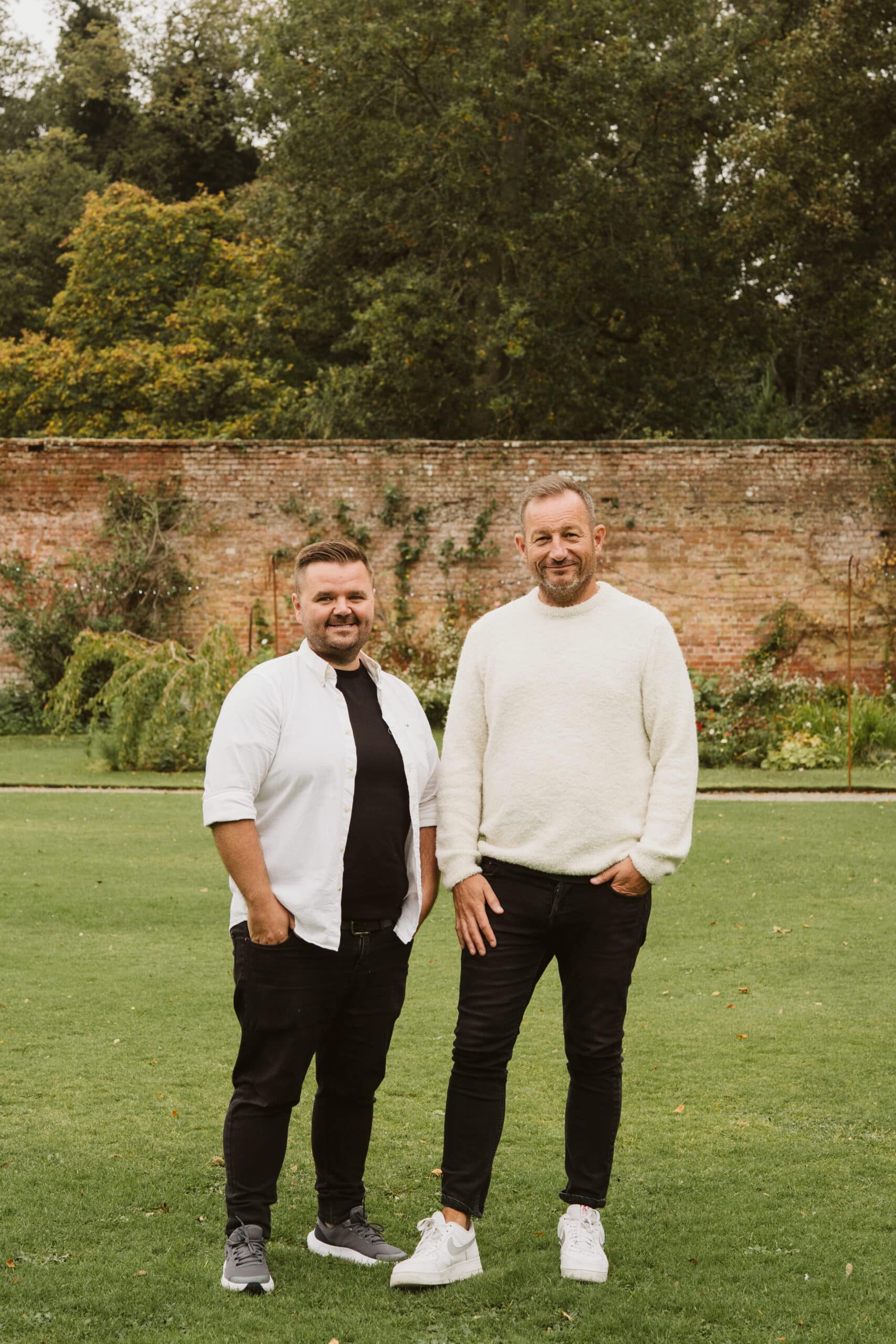 Two men standing on a lawn in front of a brick wall, surrounded by trees.