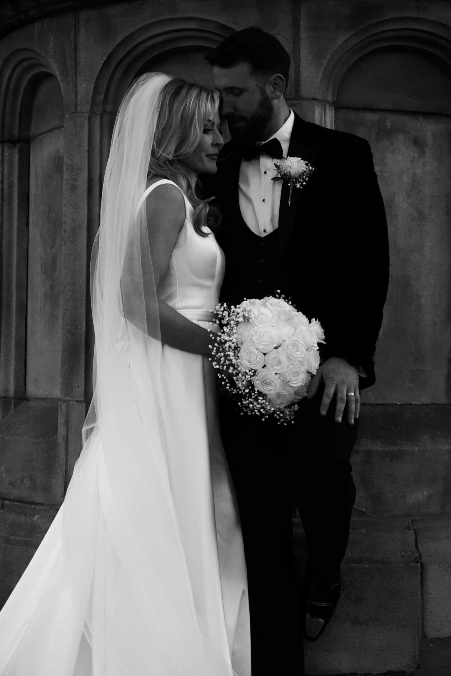 Bride and groom in formal attire, sharing a romantic moment near an ornate stone wall.
